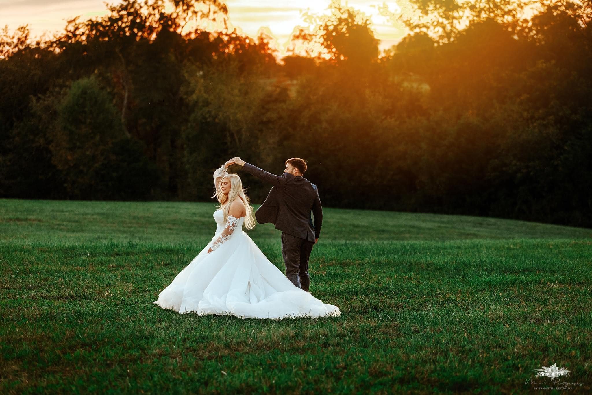 A bride and groom dancing on a grassy field during sunset, with the groom twirling the bride in her white wedding gown.