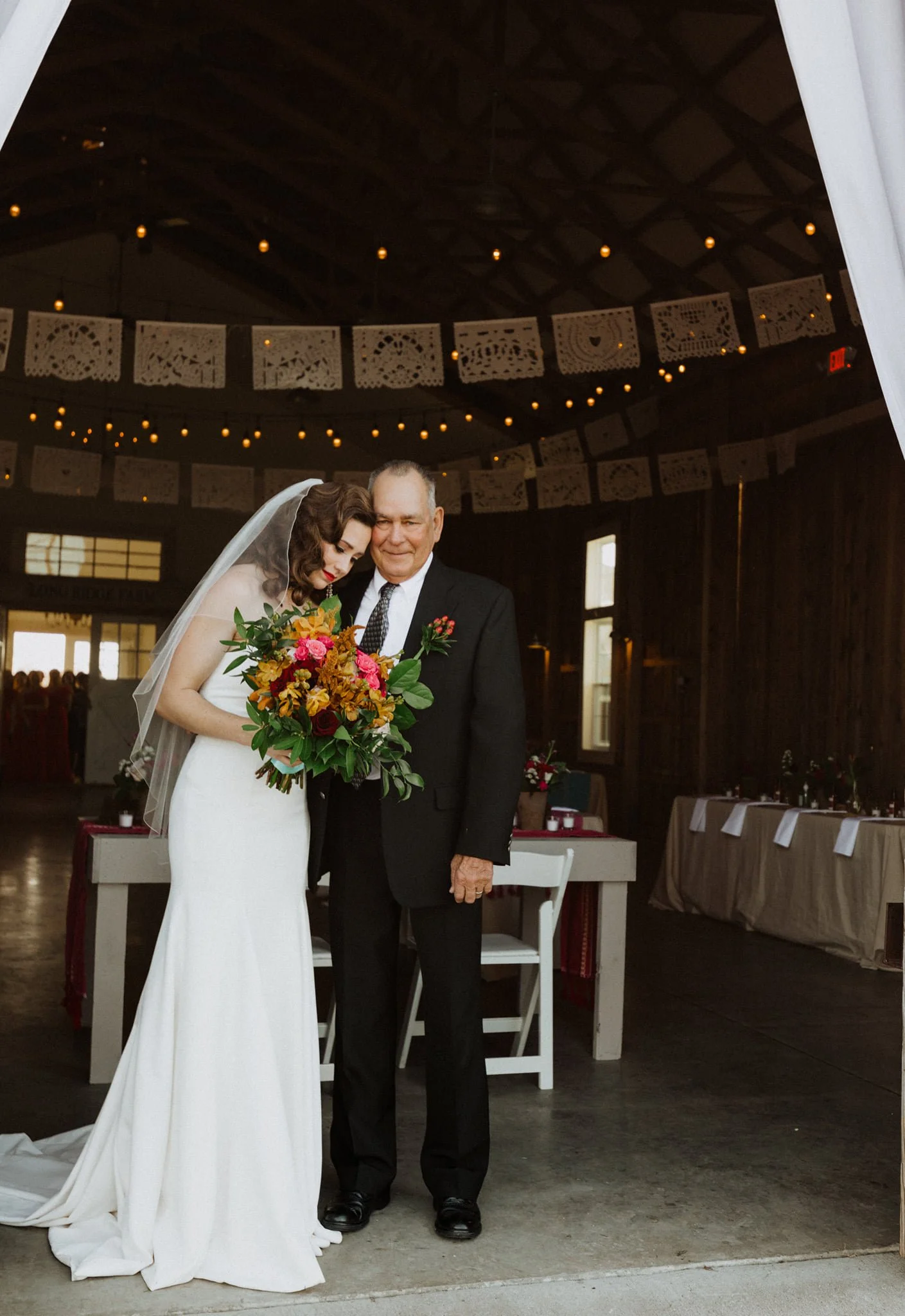 A bride in a white wedding dress holding a colorful bouquet stands next to an older man in a black suit inside a rustic indoor venue decorated with papel picado banners and string lights.