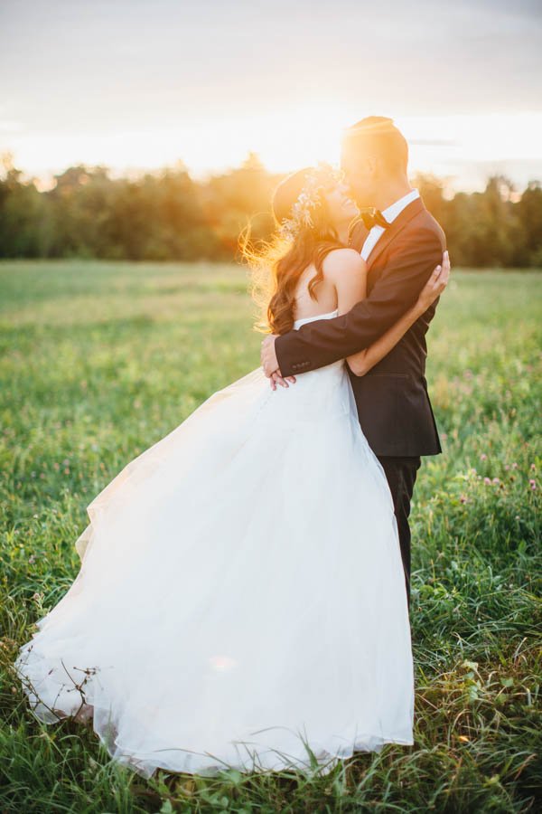 A bride and groom embracing in a grassy field at sunset, with the bride in a white wedding gown and the groom in a black tuxedo.