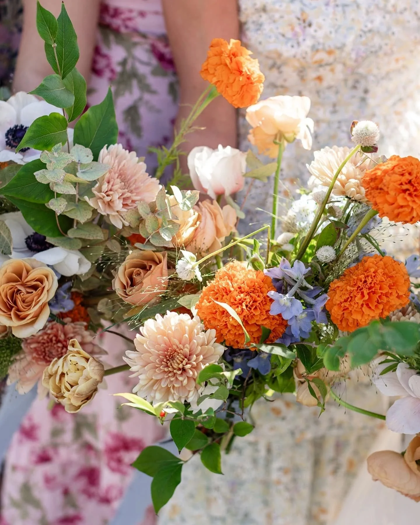 A vibrant bouquet of flowers including orange marigolds, pale pink dahlias, cream roses, white anemones with dark centers, and greenery, held by two people wearing floral dresses.