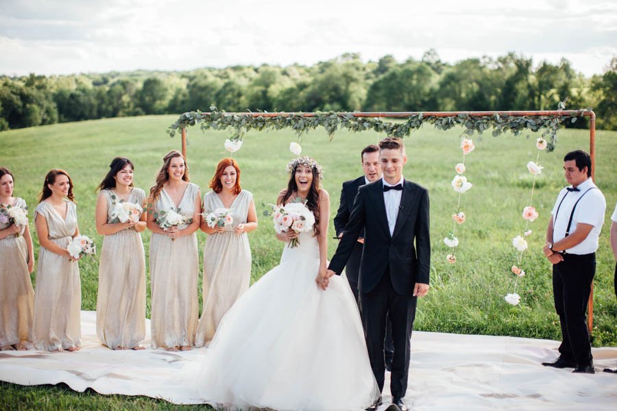 A bride and groom holding hands during an outdoor wedding ceremony on a grassy field, with bridesmaids and groomsmen standing nearby under a decorated arch with flowers and greenery.