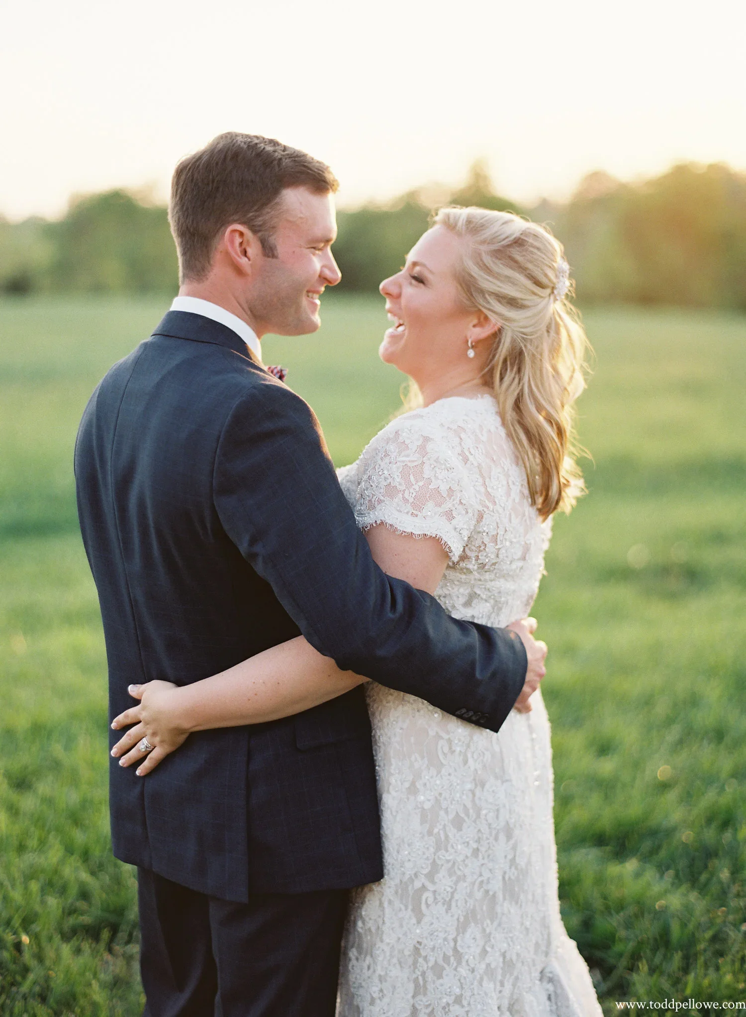 A bride and groom smiling and embracing outdoors during sunset, with greenery and trees in the background.