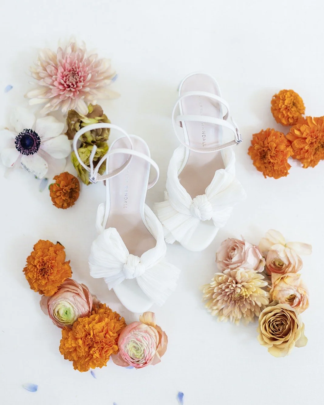 White high-heeled shoes with bow details surrounded by pink, orange, and cream flowers on a white surface.