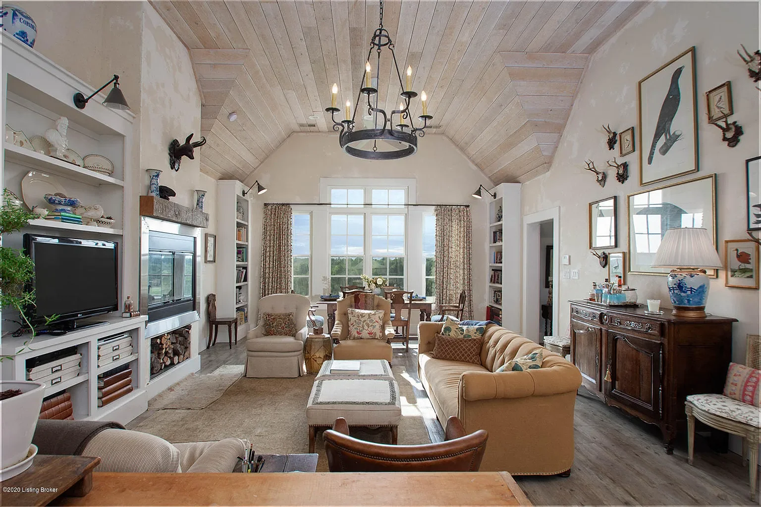 Living room with vaulted wooden ceiling, beige walls, a black chandelier, large window with curtains, and various furniture including sofas, armchairs, a wooden sideboard, and a dining table in the background. Decor includes framed art, small animal 