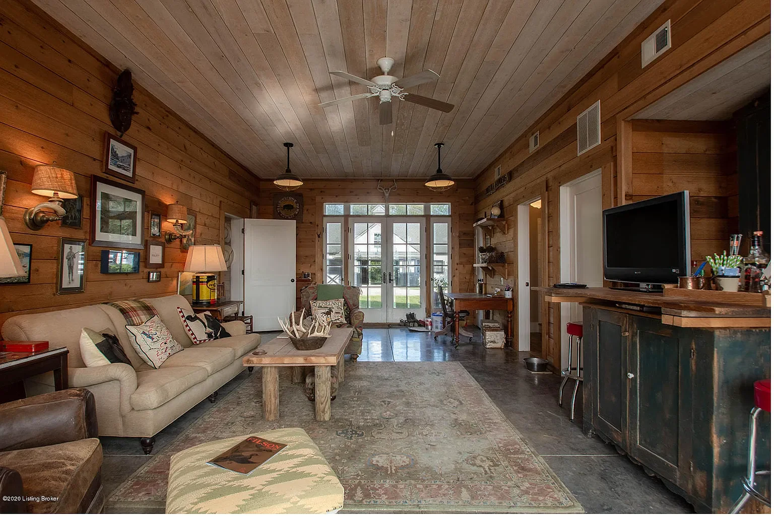 Living room with wooden walls and ceiling, furniture including a beige sofa with pillows, a wooden coffee table with antlers, a patterned ottoman, and a TV on a wooden stand. Decor includes lamps, pictures, and a mounted animal head. French doors lea