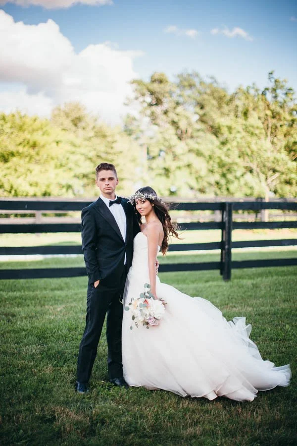 A bride and groom standing outdoors on a grassy field with trees in the background, dressed in wedding attire, with the bride holding a bouquet and wearing a floral headpiece.