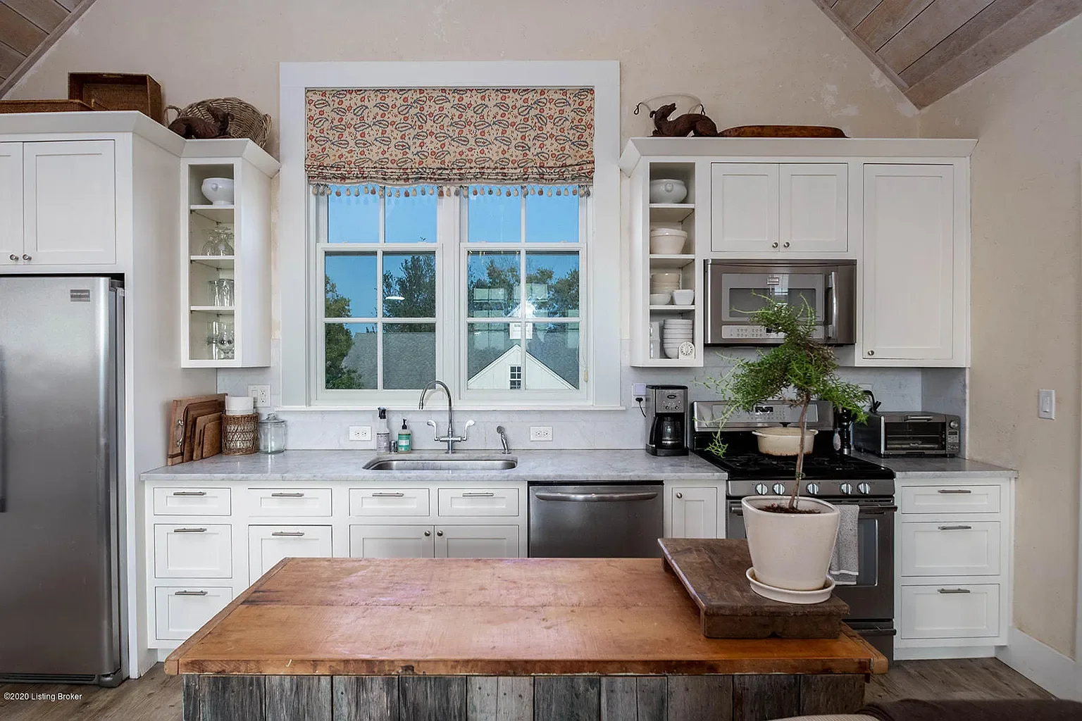 Bright kitchen with white cabinets, stainless steel appliances, and a large window with a patterned valance, featuring a wooden kitchen island with a potted plant on top.