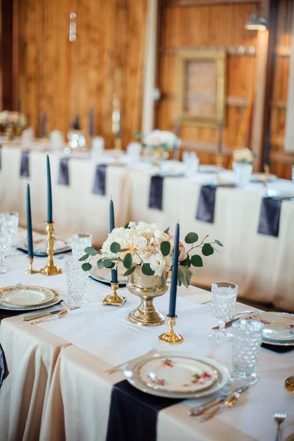 Elegant dining table with white tablecloth, gold accents, floral centerpiece, navy blue candles, and fine china in a rustic wooden room.