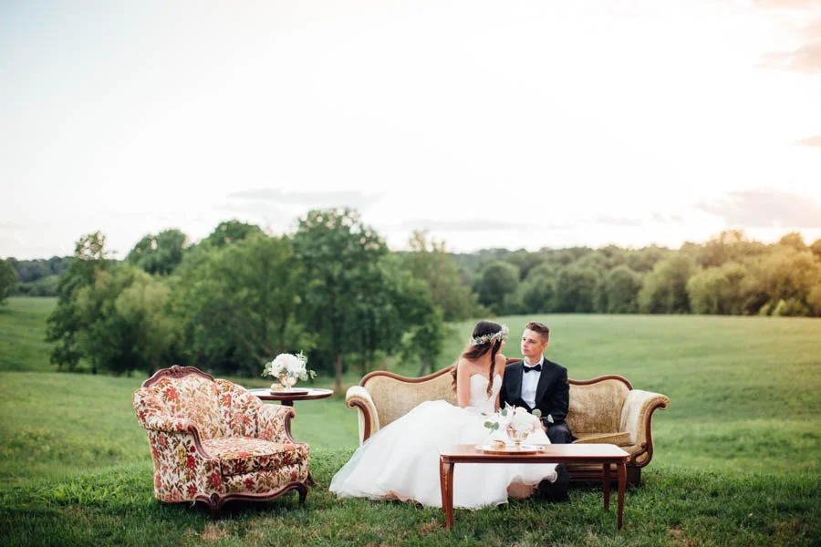 Bride and groom sitting on a vintage sofa outdoors, with a small table beside them holding flowers, set in a green field with trees in the background during sunset.
