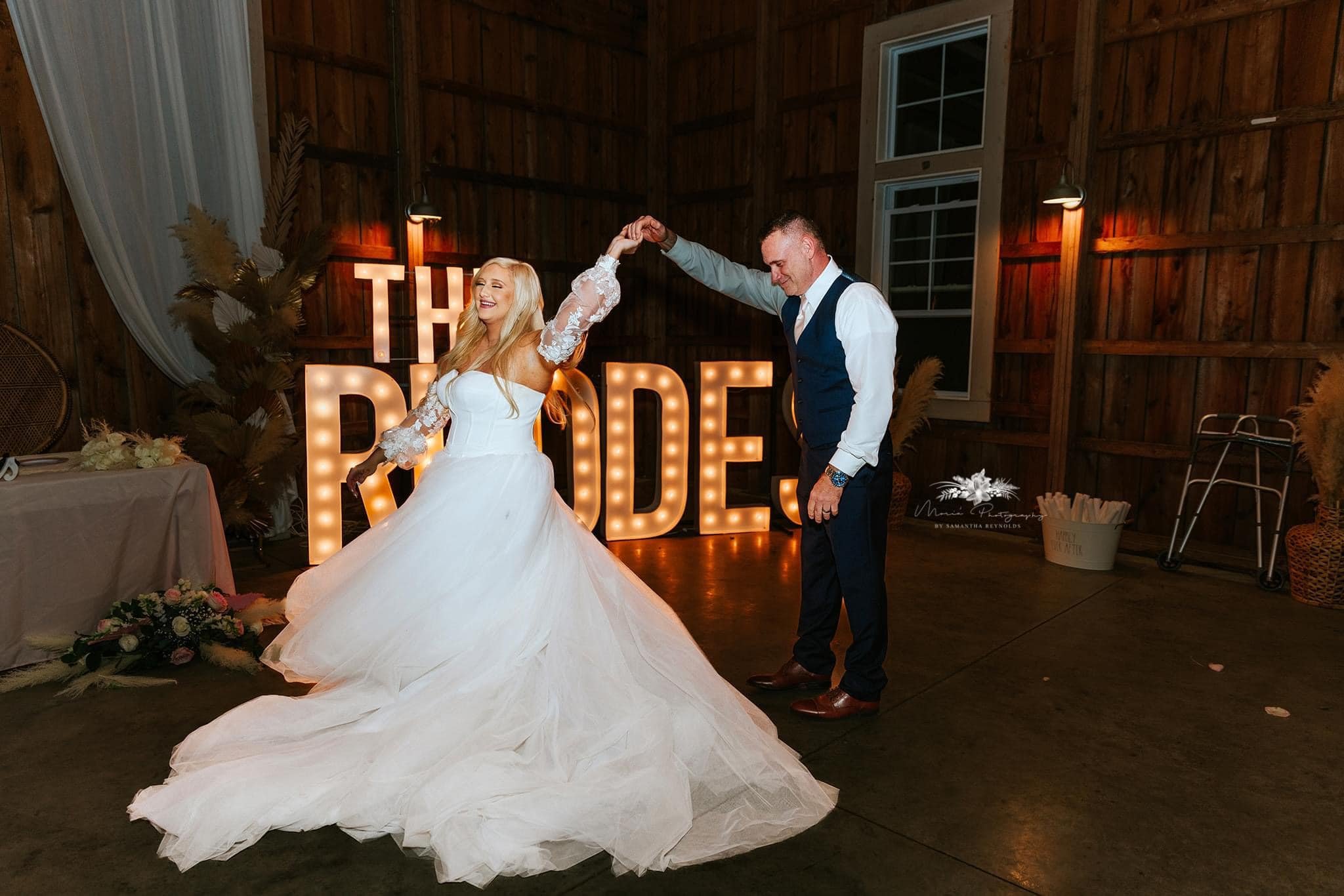 A bride and groom dancing at their wedding reception in a rustic barn. The bride is wearing a white gown with lace sleeves, and the groom is dressed in a white shirt with a vest and tie. In the background, illuminated marquee letters spell out 'THE BRIDE'.