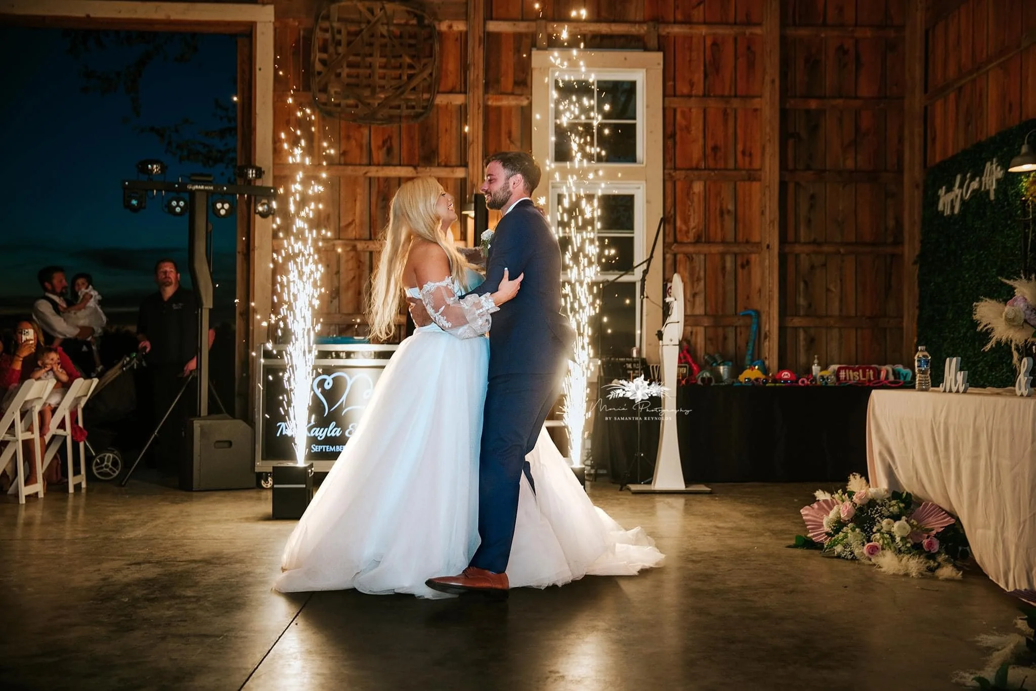 A bride and groom dancing at their wedding reception surrounded by sparklers, with guests watching in the background, inside a rustic wooden venue.