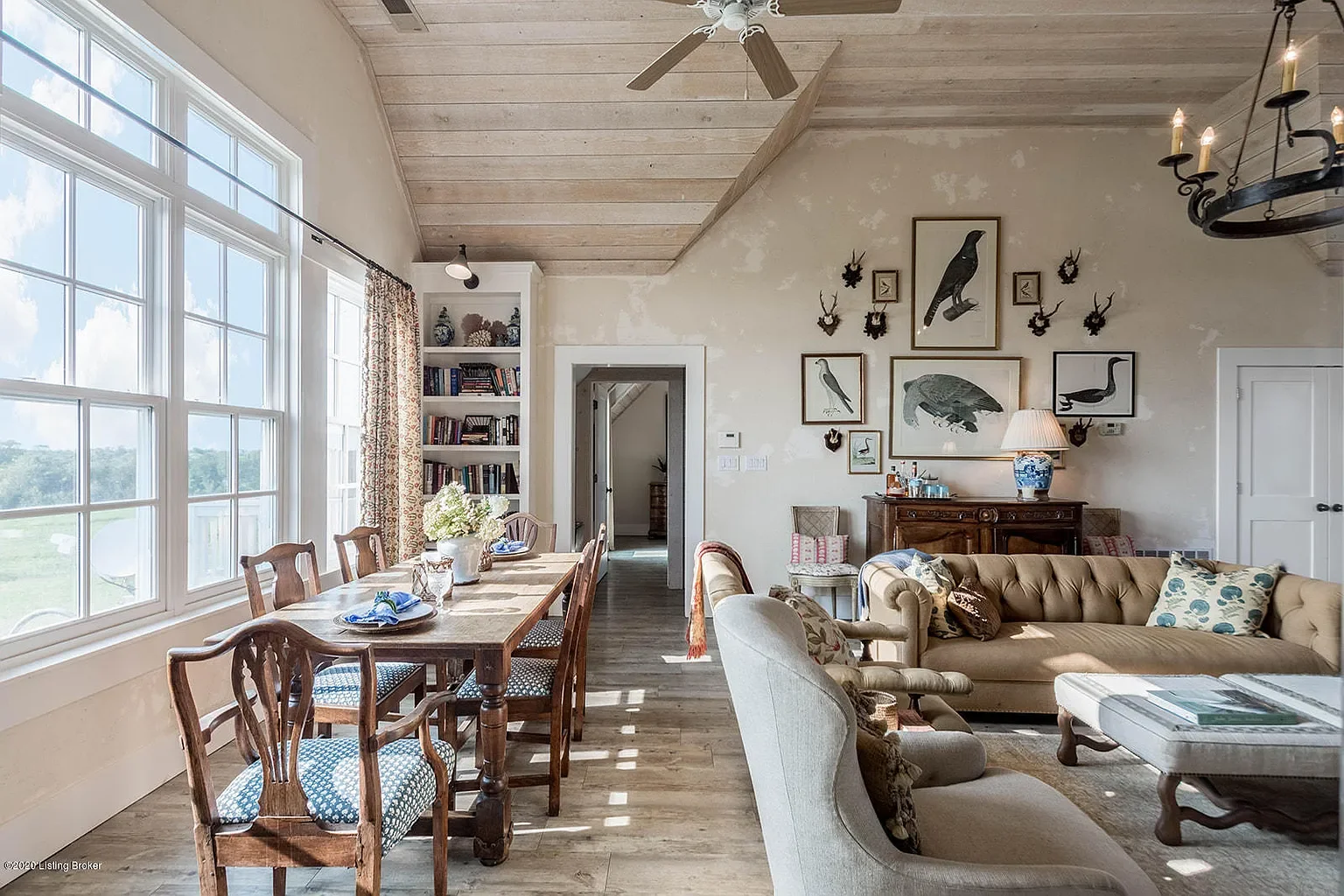 Bright living room with large windows, wooden ceiling, dining area with wooden table and chairs, and a sitting area with a beige sofa, armchairs, and various framed bird and animal artwork on the wall.