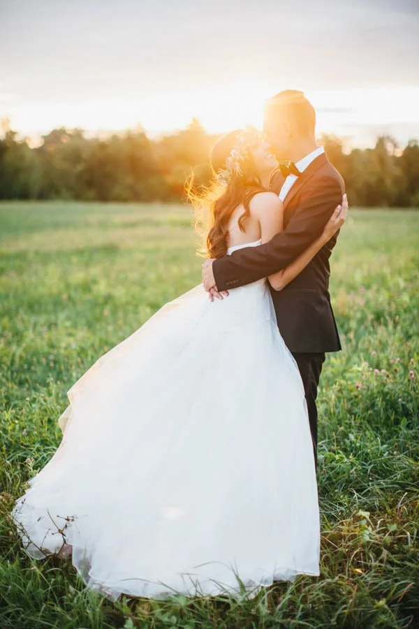 A bride and groom embracing in a grassy field at sunset, with the bride wearing a white gown and the groom in a black suit.