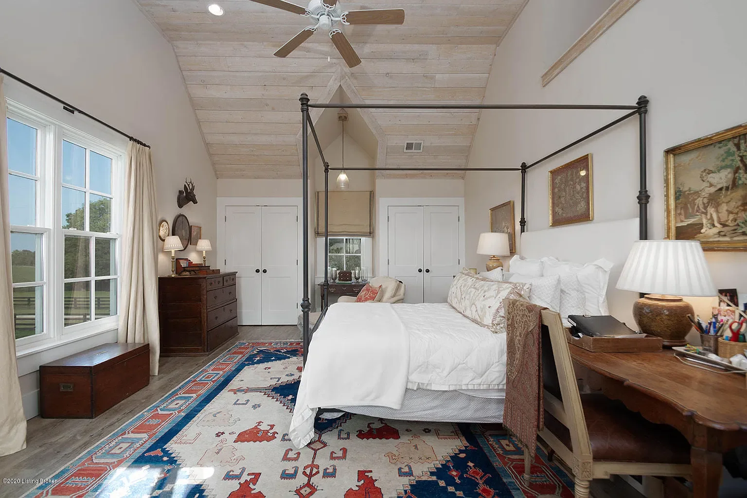 A bedroom with a vaulted wooden ceiling, large window with curtains, four-poster bed with white bedding, wooden dresser with lamps, colorful patterned area rug, and artwork on the walls.