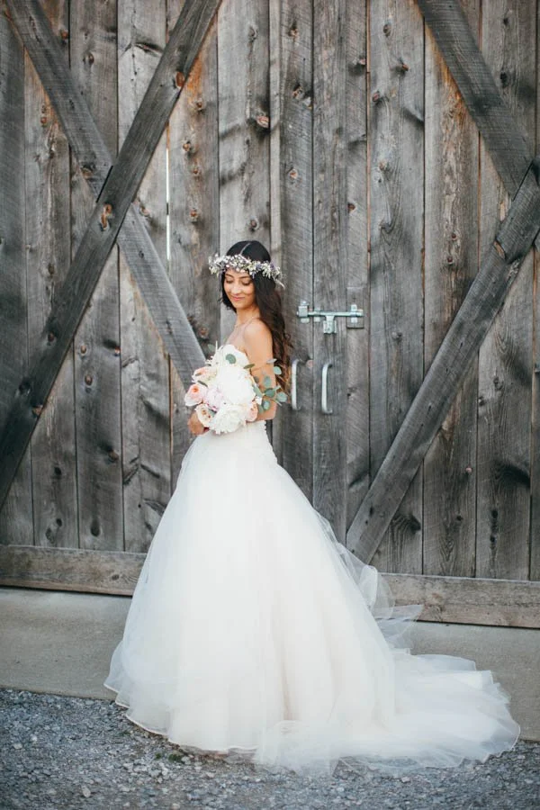 Young woman in a white wedding dress holding a bouquet, standing in front of a rustic wooden barn door, wearing a floral crown.