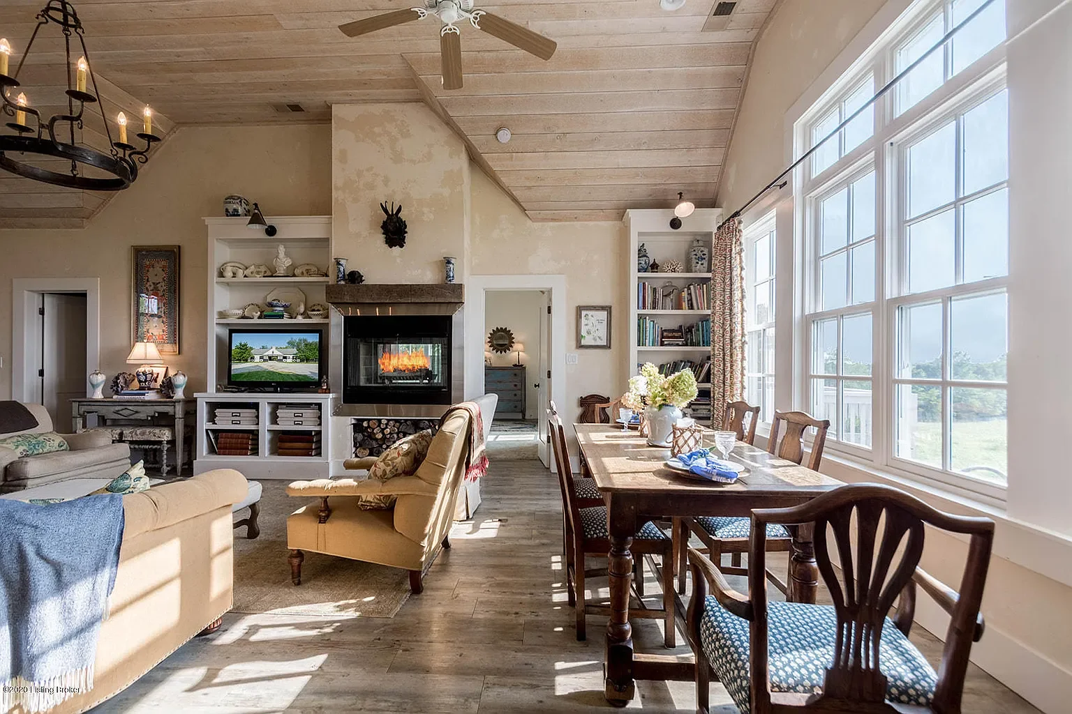 Living and dining room with large windows, wooden floors, and a stone fireplace with a fire. Furniture includes a beige sofa, a yellow armchair, and a dark wooden dining table with chairs. Decor features bookshelves, a chandelier, and a ceiling fan.