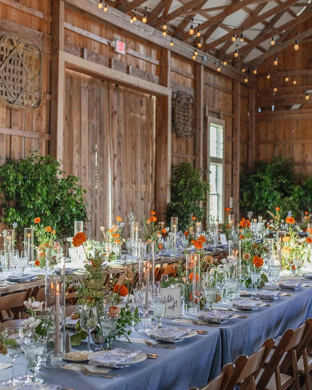 Long wooden banquet table decorated with pink and orange floral arrangements, glassware, candles, and table settings, inside a rustic barn with string lights hanging from the ceiling and green plants in the background.