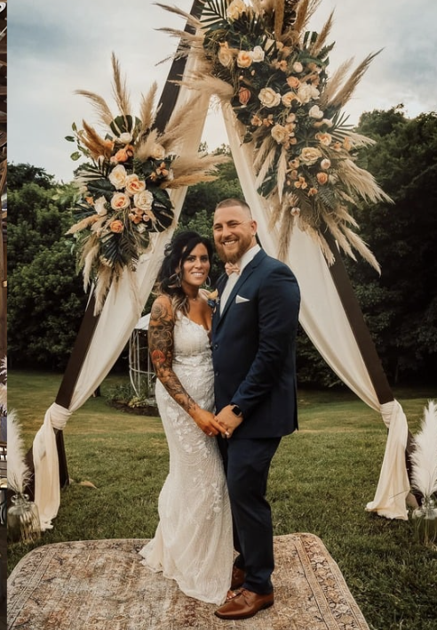 A bride and groom standing hand in hand outdoors in front of a floral wedding arch on a grassy lawn, with trees in the background. The bride is in a white lace wedding dress with tattoos on her arms, and the groom is in a navy blue suit with a white shirt and brown shoes. They are smiling after their wedding ceremony.