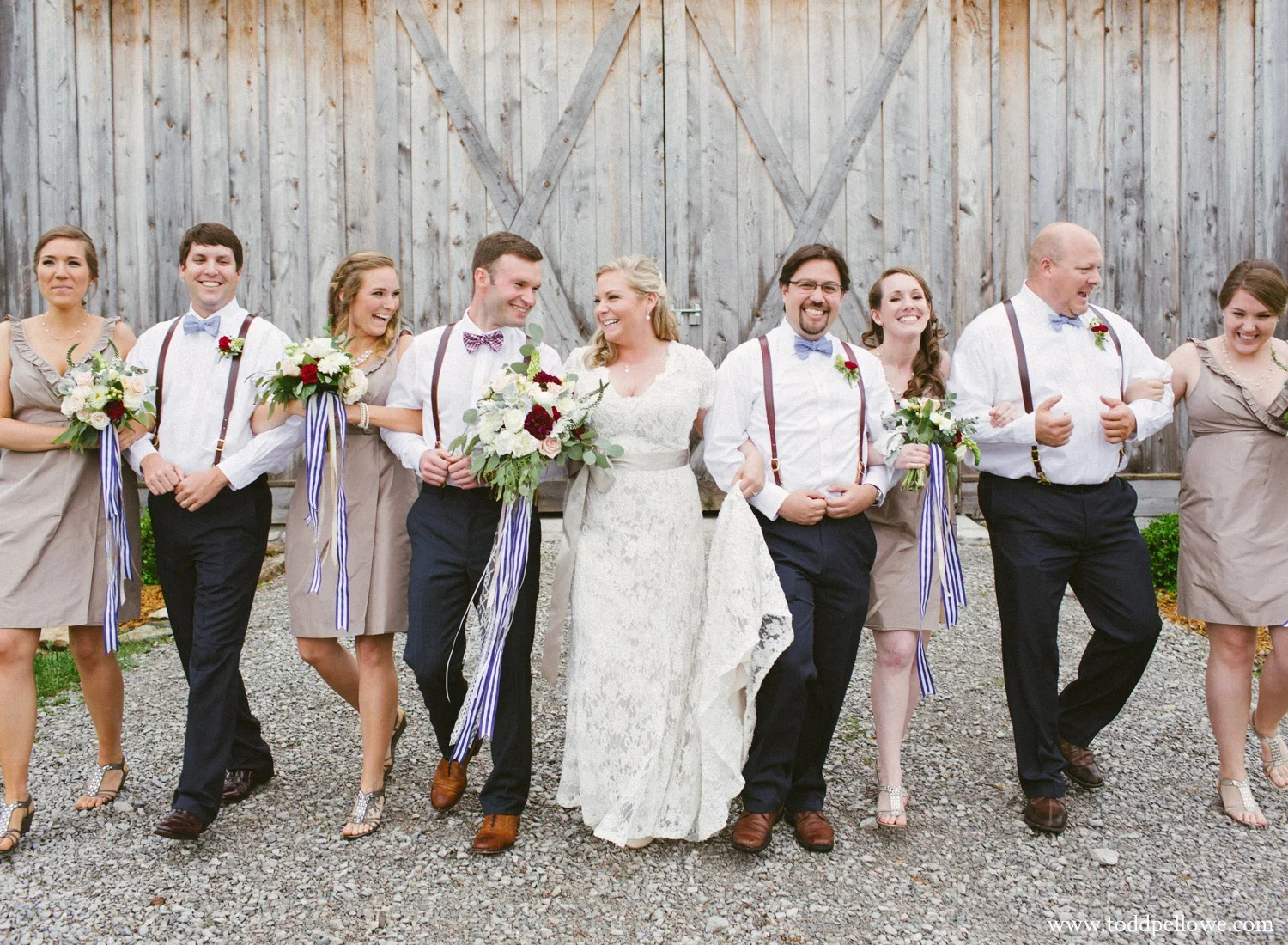 A group of nine people, including a bride and groom, walking arm-in-arm outdoors in front of a wooden barn, all smiling and holding bouquets.