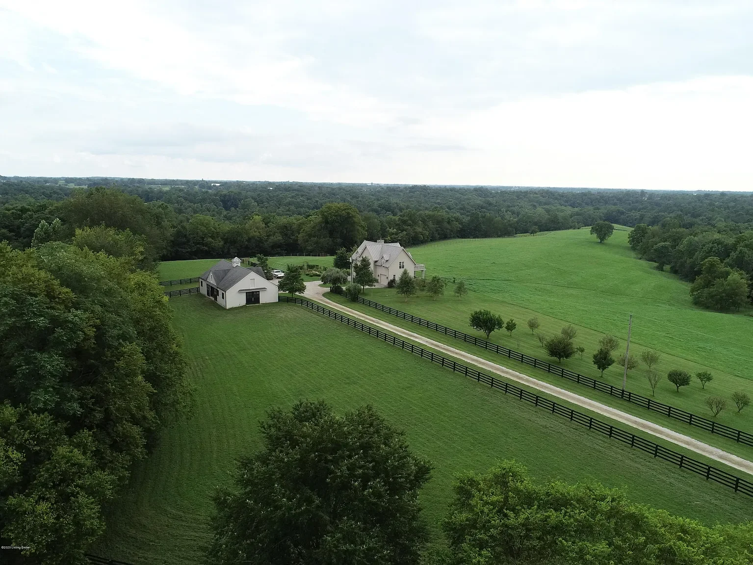 An aerial view of a rural farm with white buildings, a driveway, green fields, and trees under a cloudy sky.