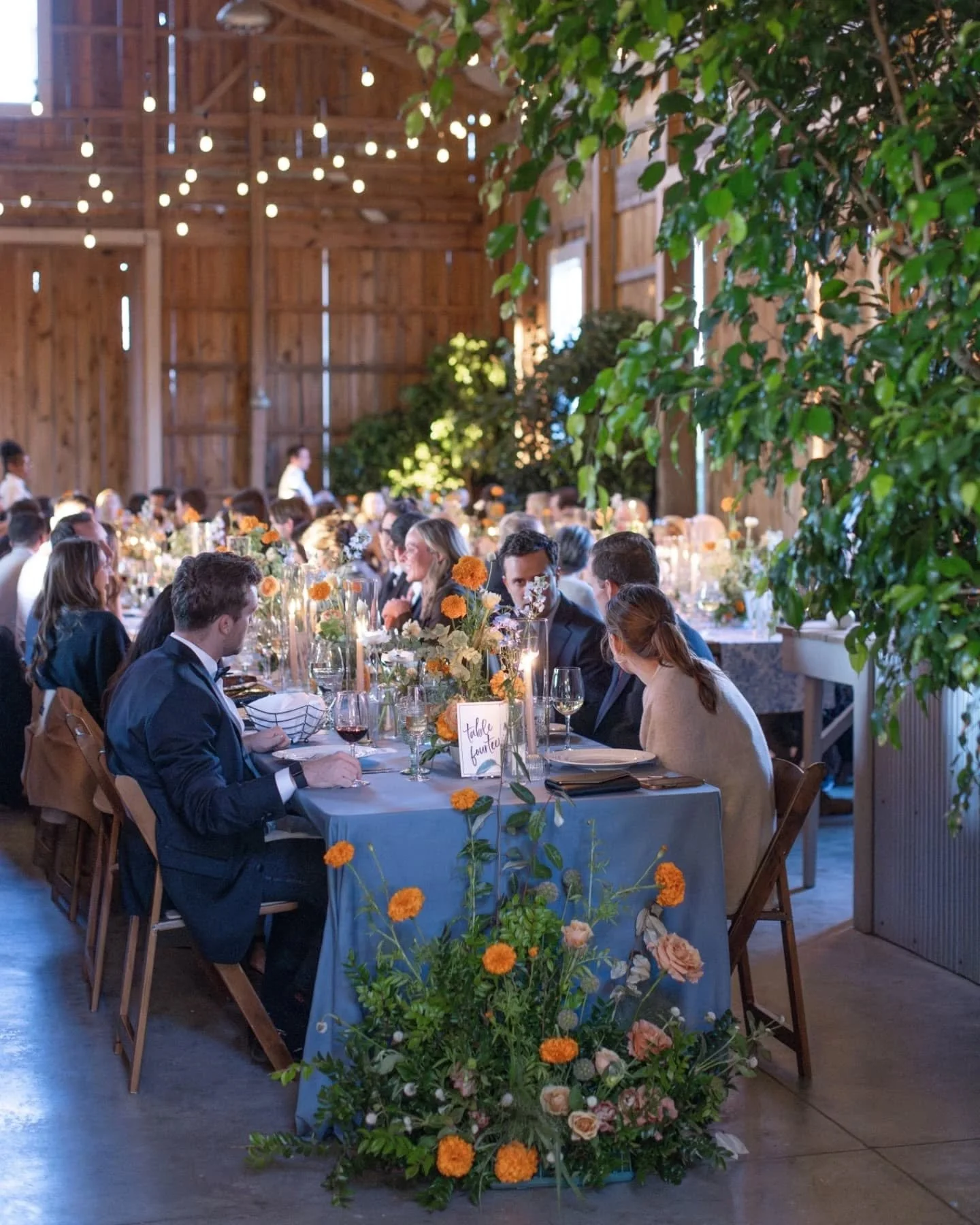 Indoor wedding reception with long tables decorated with flowers and candles, guests dressed formally, in a rustic barn with string lights and greenery.