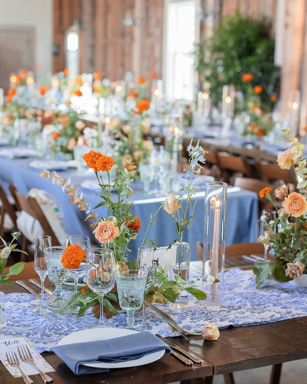 Decorated banquet table set with floral centerpieces, candles, glassware, silverware, and blue napkins, with a rustic wooden wall and large windows in the background.
