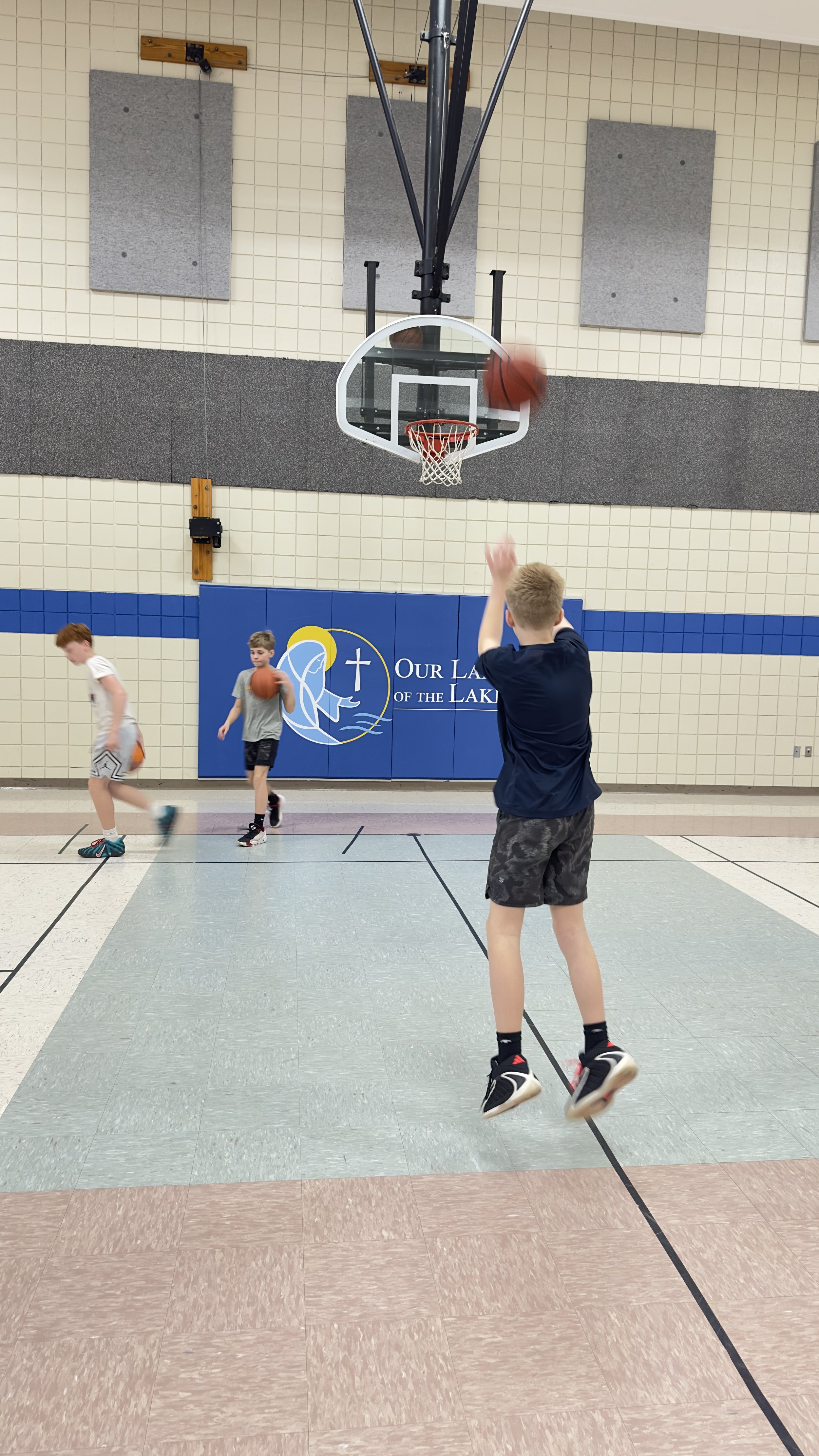 Children playing basketball in an indoor gym, with one boy shooting the ball towards the hoop and others practicing with basketballs. The gym has tiled walls and a blue sign that reads 'Our Lady of the Lake'.