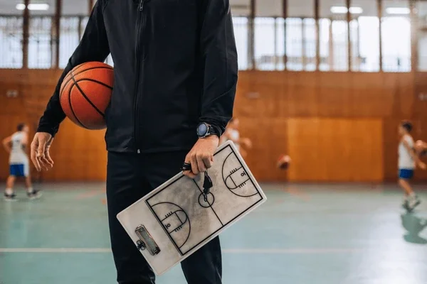Person holding a basketball and a clipboard with a basketball court diagram, in a gymnasium with other players in the background.