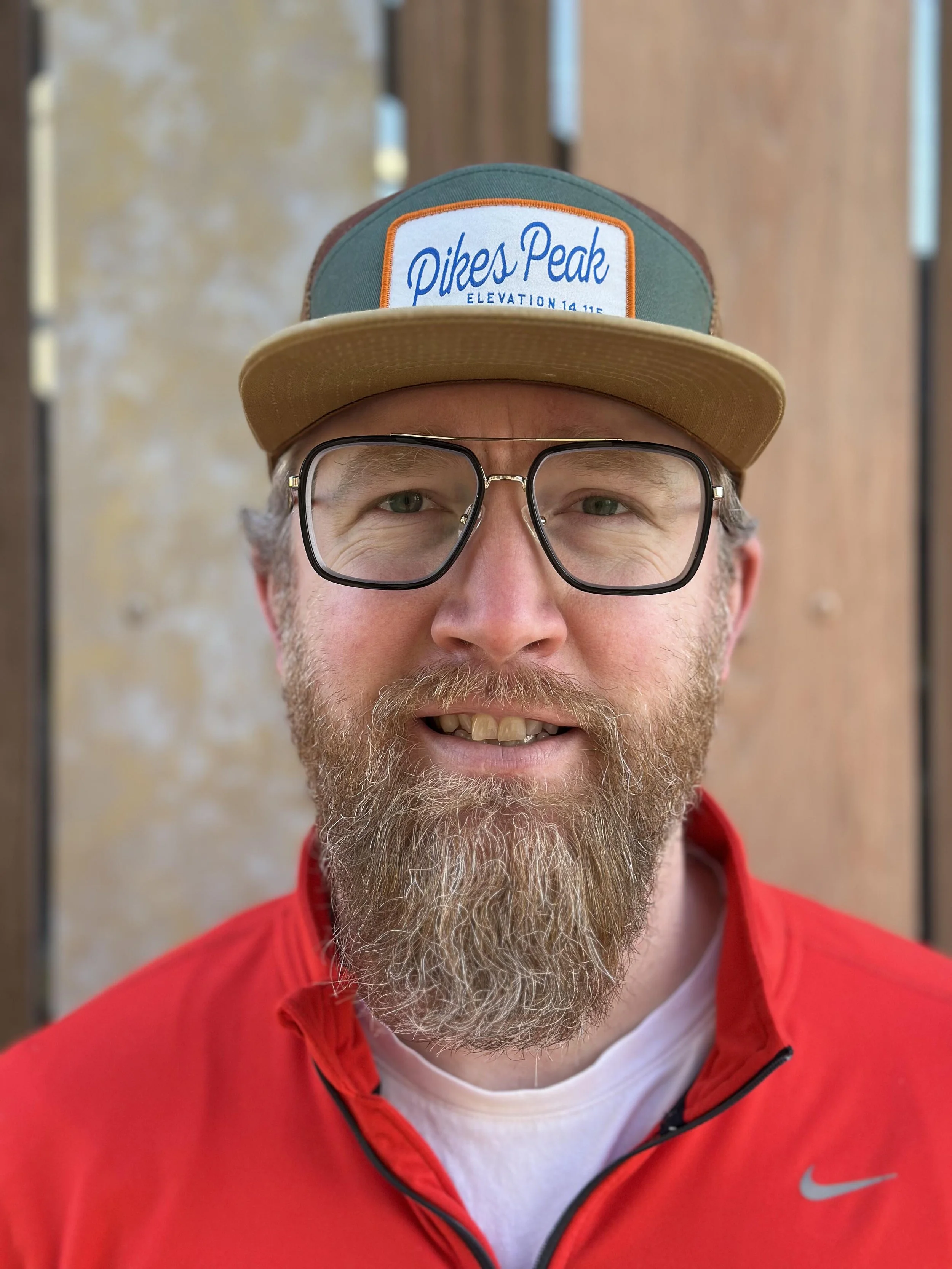 A bearded man wearing glasses, a red jacket, and a cap with 'Pikes Peak' and 'Elevation 14,115' written on it, smiling in front of a wooden and stone background.