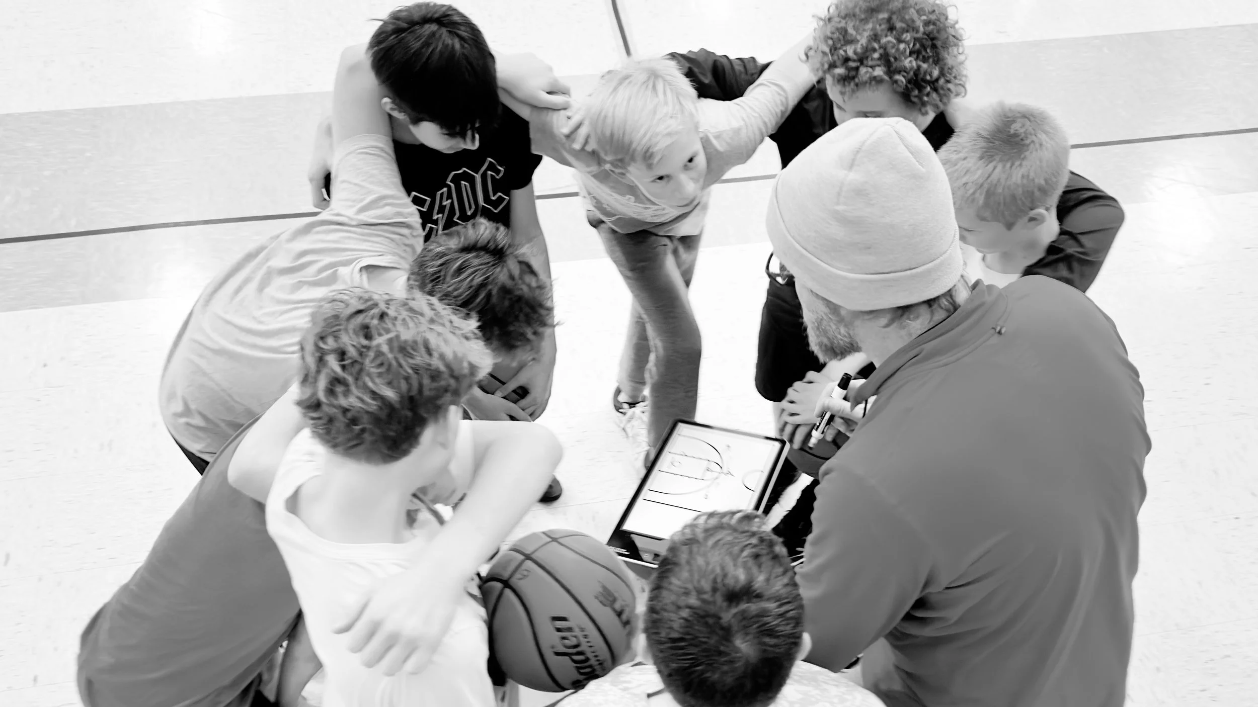 A group of children gathered in a circle around an adult coach with a tablet, watching a basketball game diagram, inside a gymnasium.