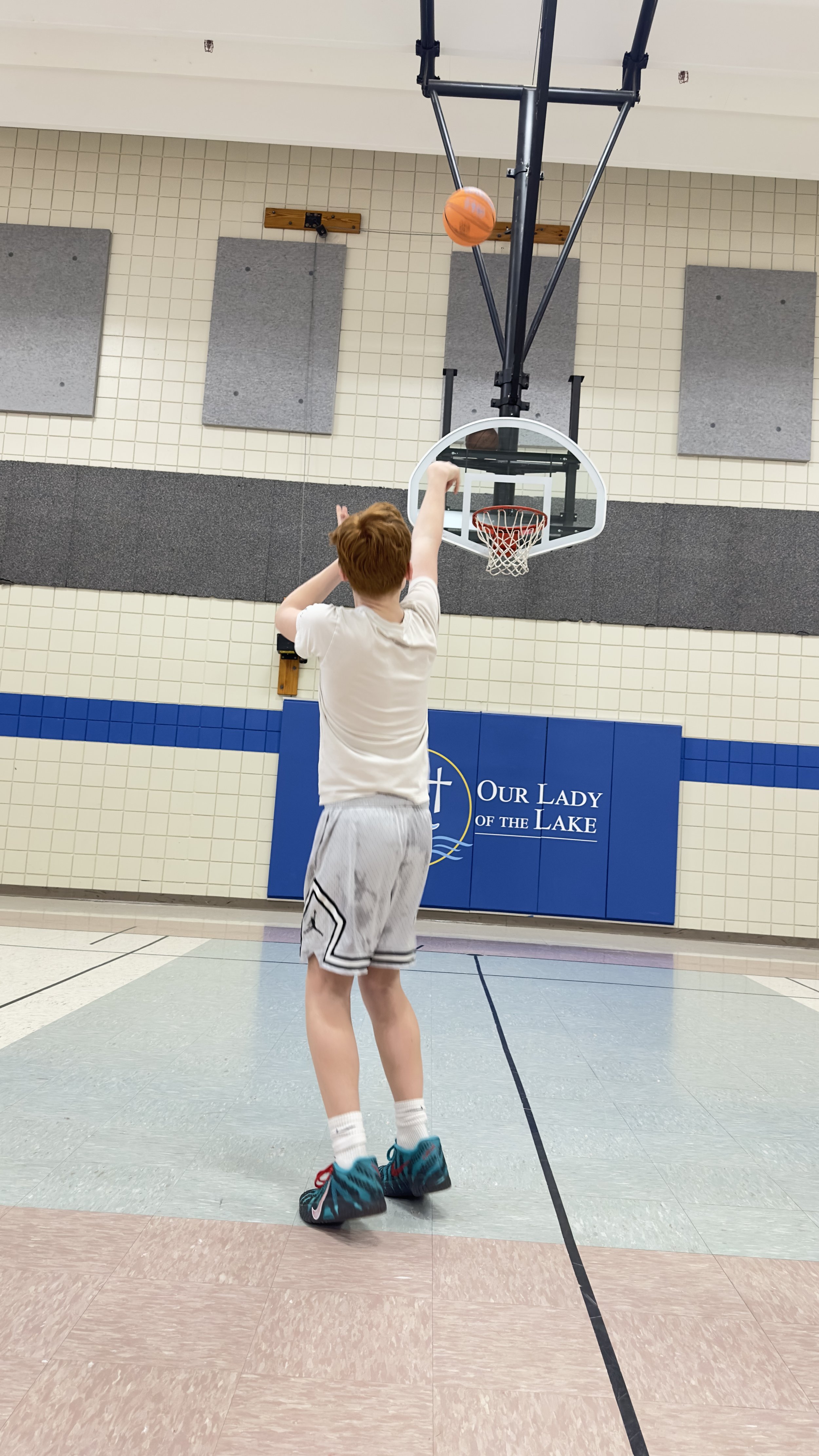 A boy with red hair shooting a basketball towards a hoop in a gymnasium. The gym has beige tiled walls, a blue padded section with the logo 'Our Lady of the Lake', and gray panels on the wall.