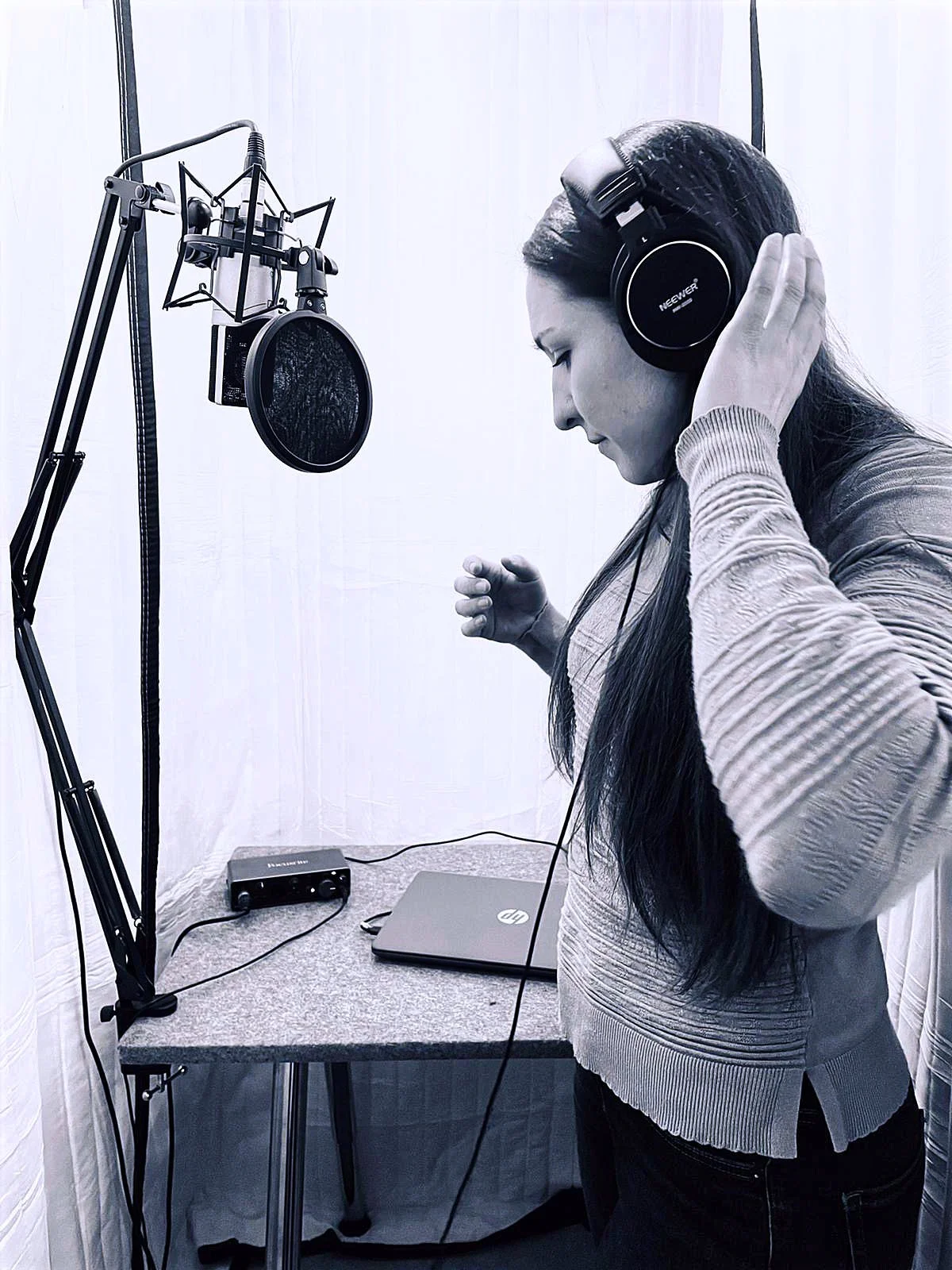 A woman in a recording booth wearing headphones, standing near a microphone on a stand, with a laptop on a table beside her.