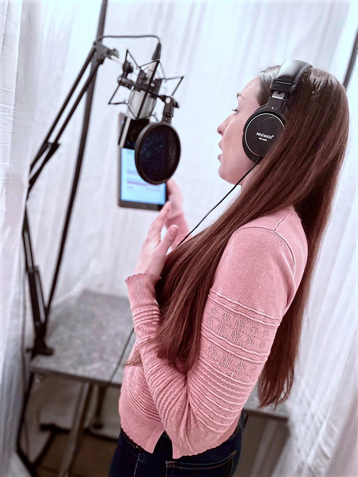 A woman with long reddish-brown hair recording vocals in a soundproof studio, wearing large headphones, holding a smartphone in one hand, and standing in front of a professional microphone.