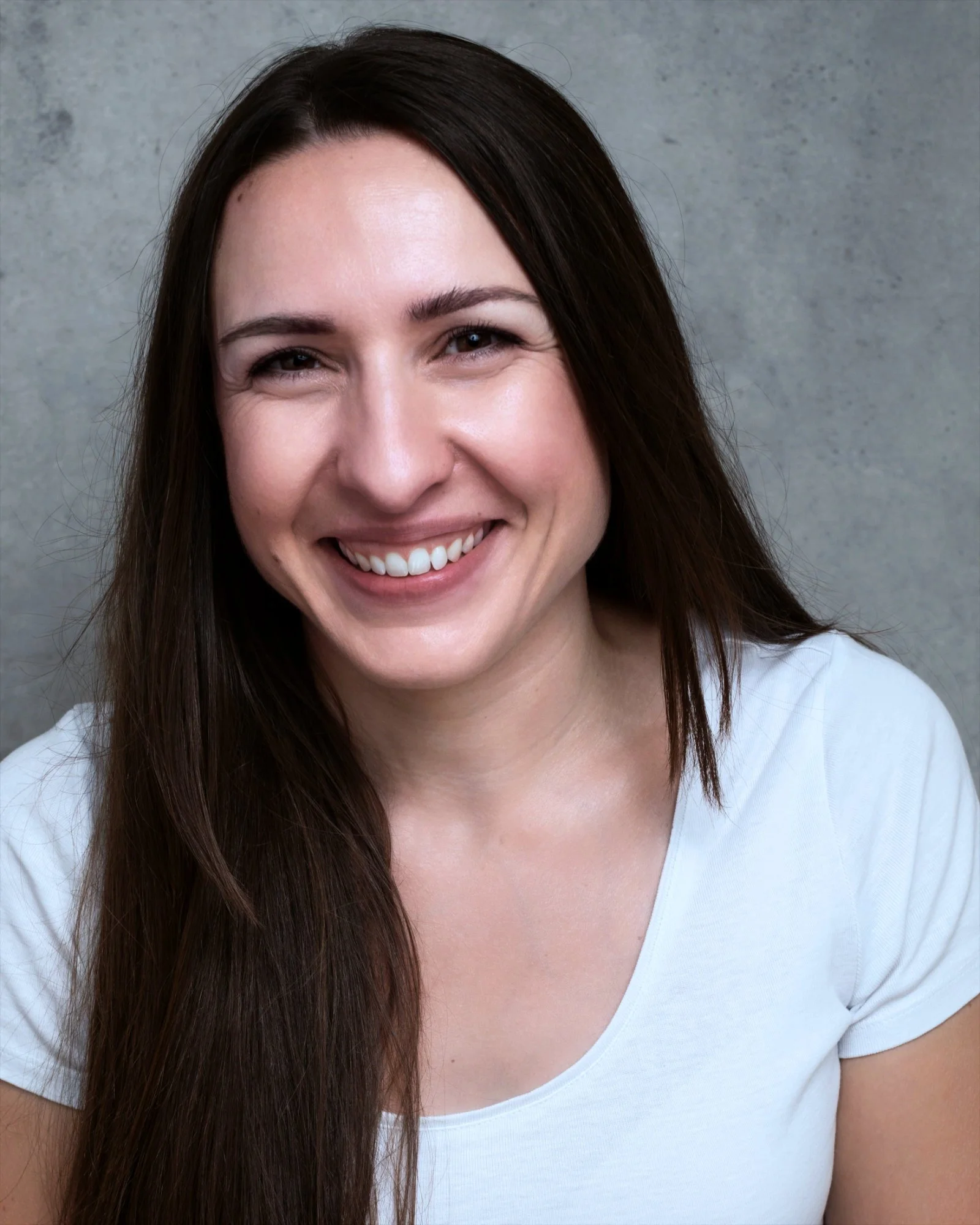 A woman with long dark brown hair smiling, wearing a white t-shirt, standing against a gray textured background.