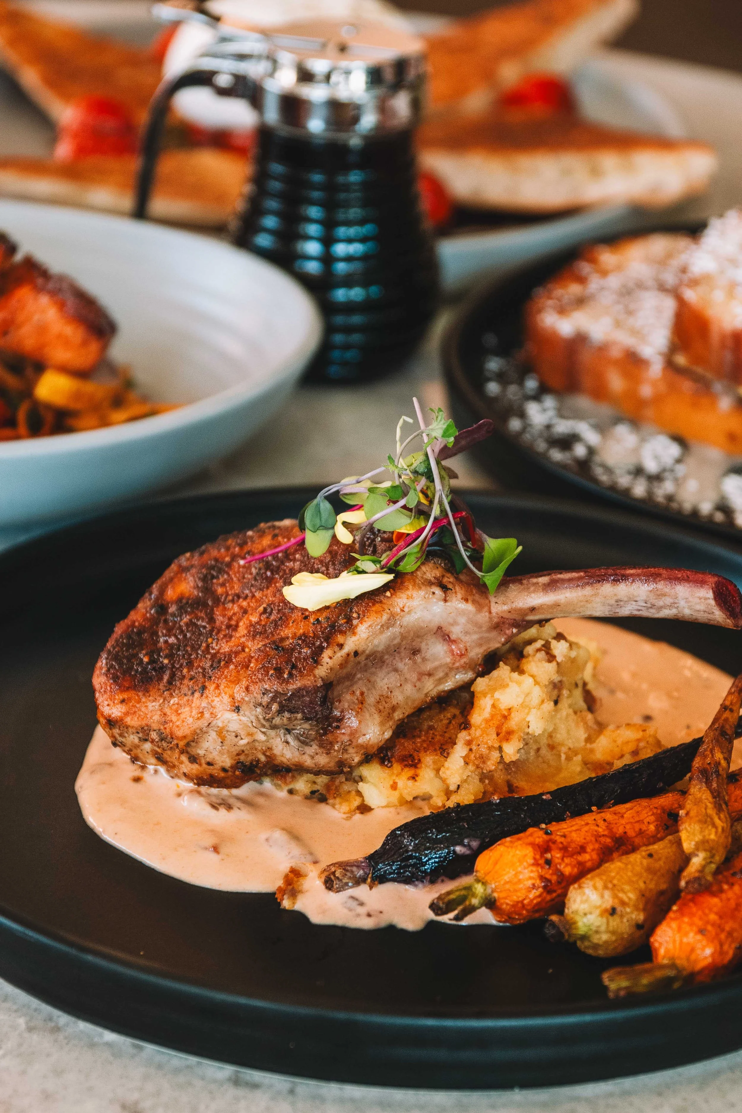Close-up of a roasted pork chop garnished with microgreens, served with roasted carrots on a black plate, cream sauce underneath, with other dishes in the background.