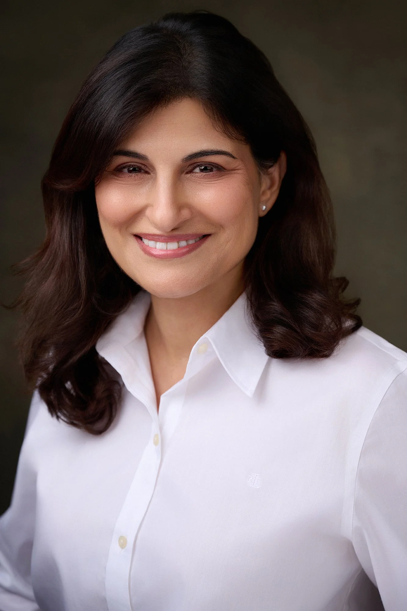 A woman with shoulder-length dark brown hair smiling, wearing a white button-up shirt, against a neutral background.