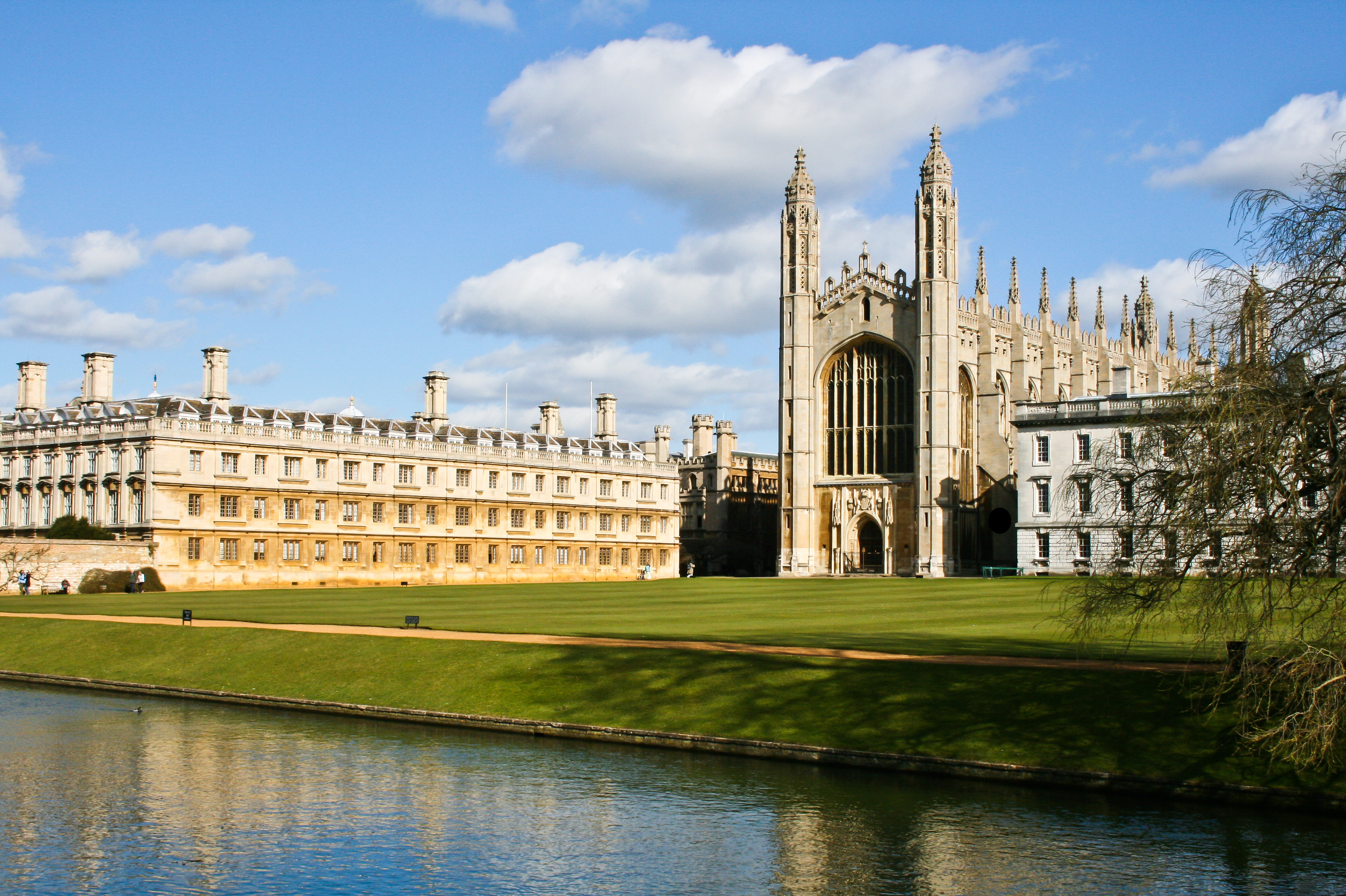 Photograph of a historic university building with Gothic architecture, situated near a river with a grassy area in the foreground, under a partly cloudy sky.