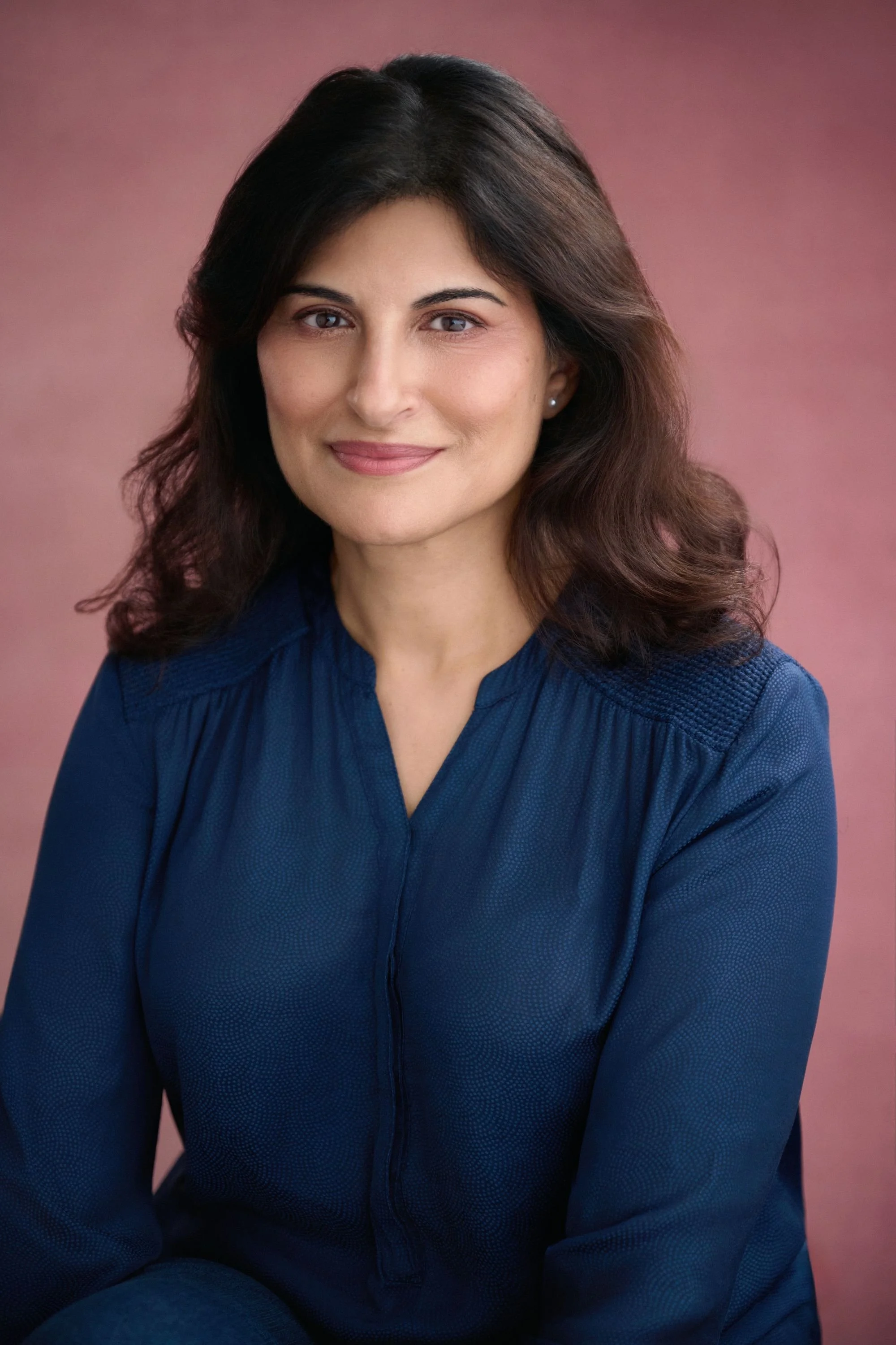 A woman with dark brown hair wearing a blue blouse, smiling softly, against a pinkish background.