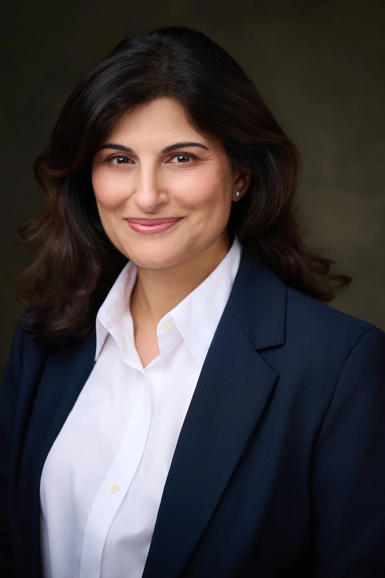 Professional woman with dark wavy hair wearing a white shirt and navy blazer, smiling against a dark background.