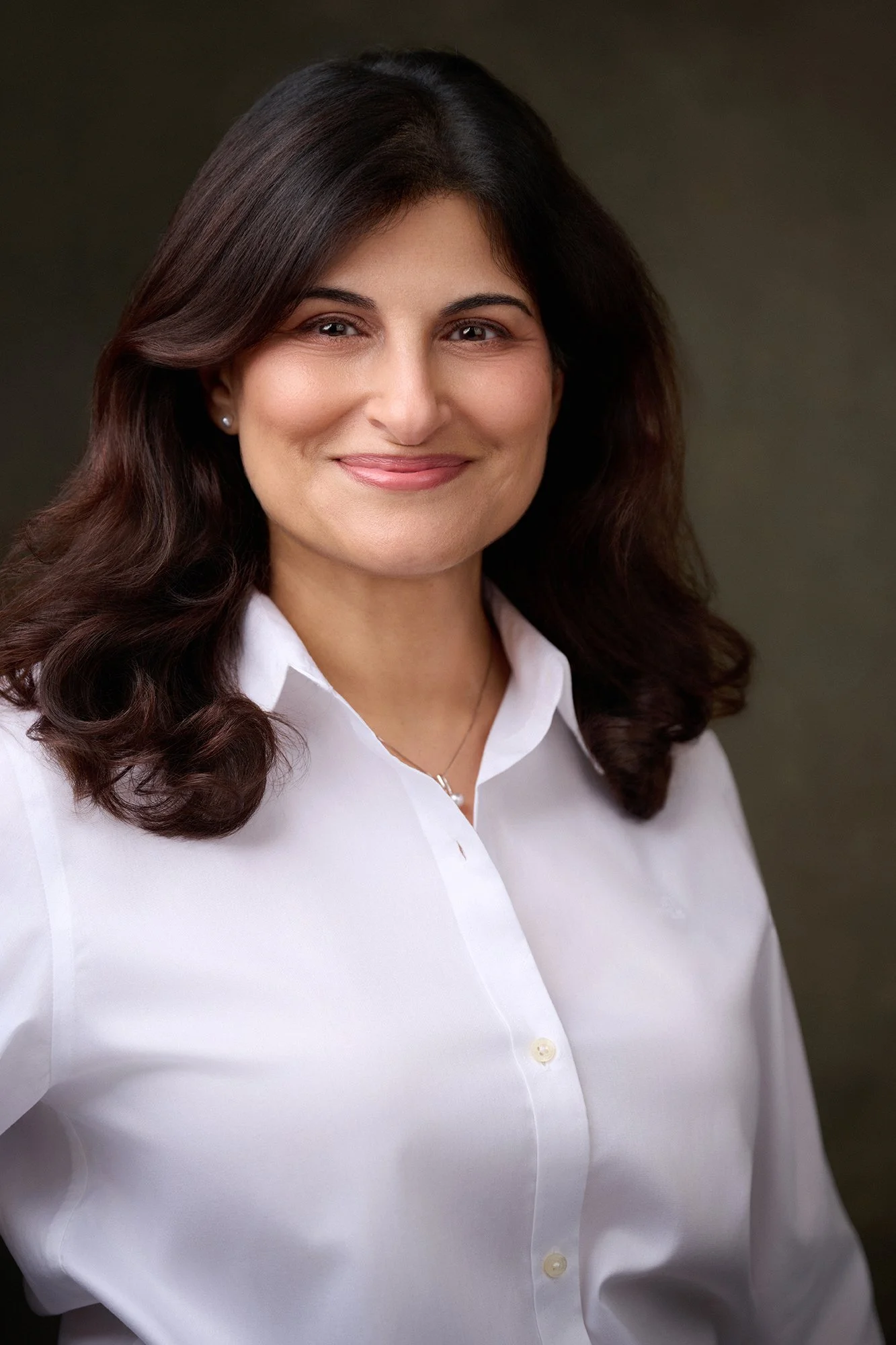 A woman with dark, wavy hair wearing a white collared shirt and a delicate necklace, smiling against a dark background.