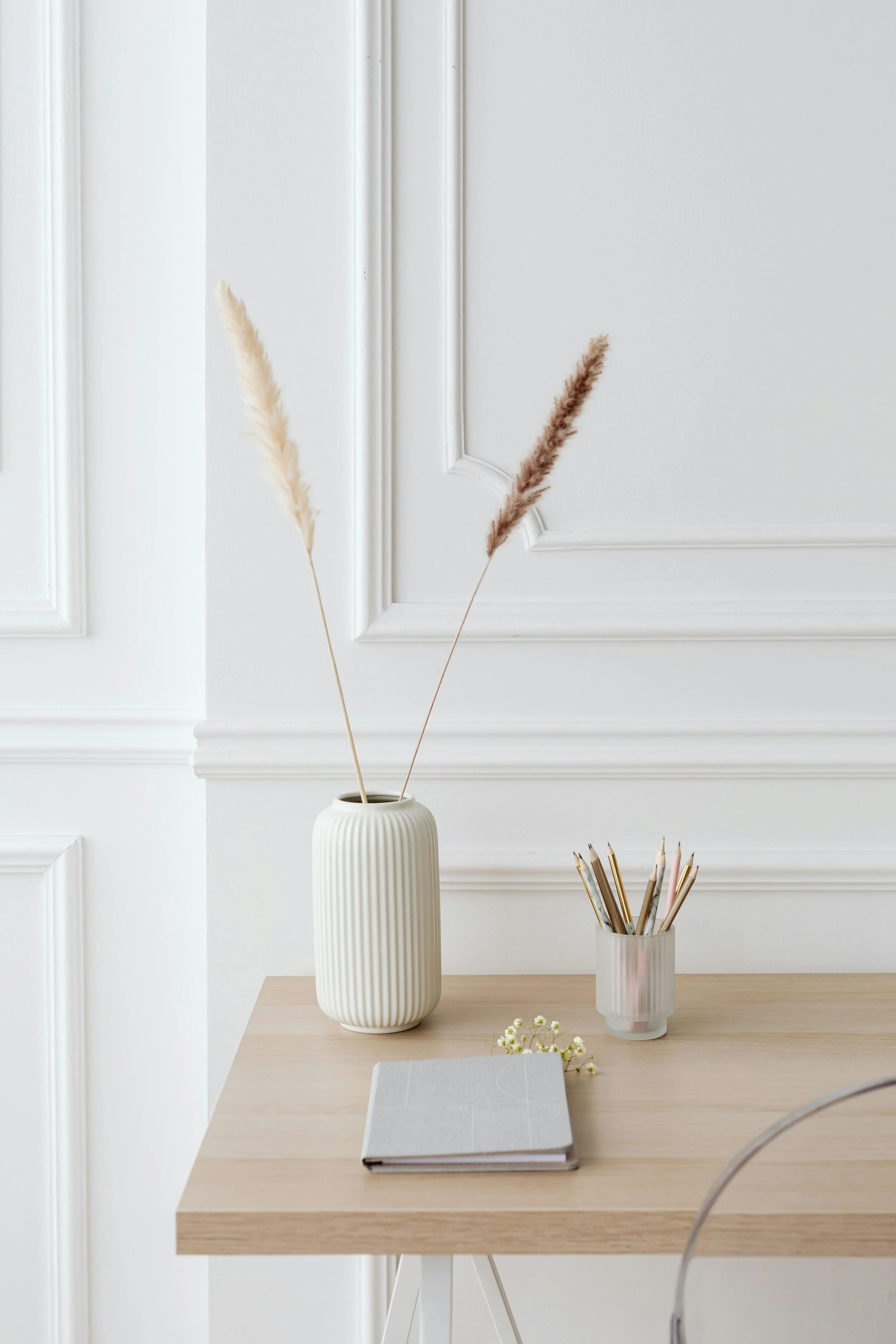 Minimalist desk with a wooden top, white vase with pampas grass, a small container with colored pencils, and a closed notebook, set against a white wall with decorative molding.