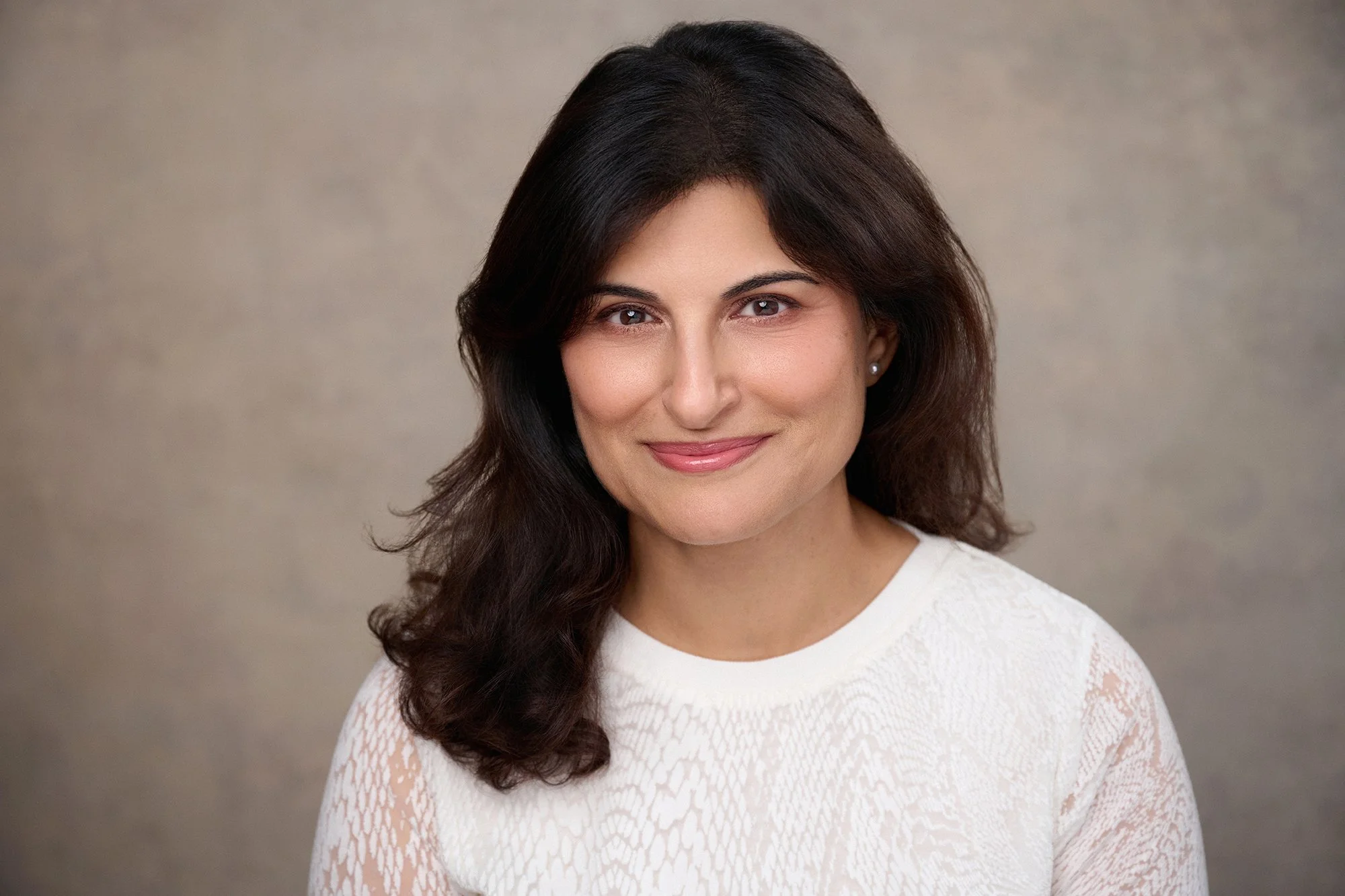 A woman with dark brown hair styled in loose waves, wearing a white textured top, smiling softly, against a neutral beige background.