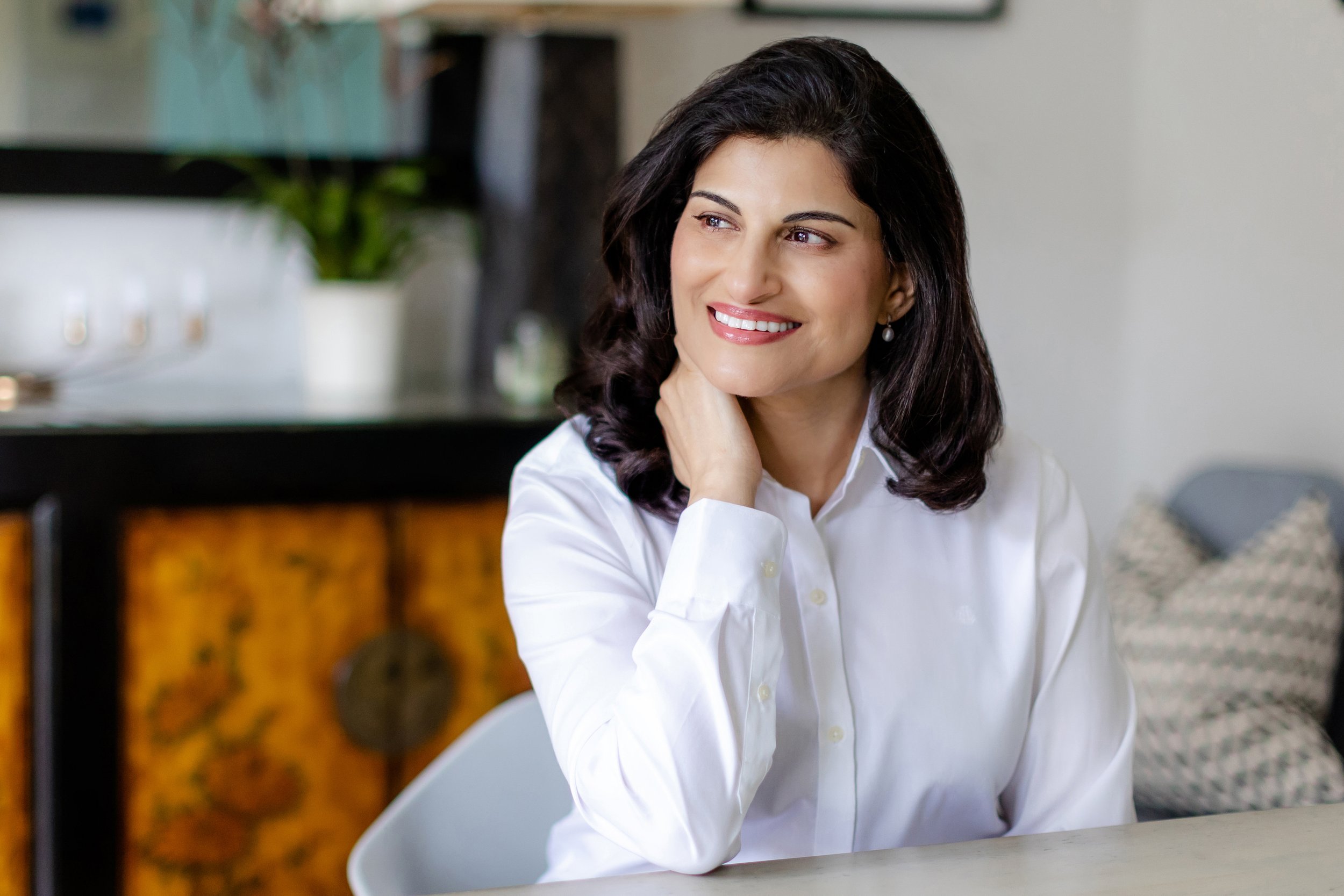 A woman with dark, wavy hair and wearing a white blouse sitting at a table with a smile, in a modern indoor setting.