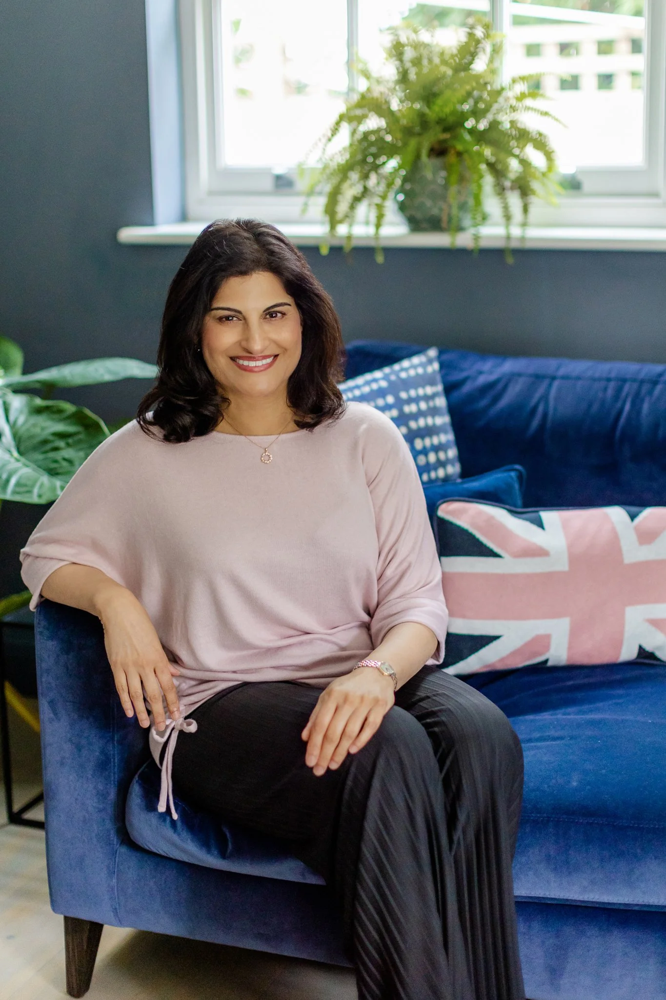 A woman with dark hair sitting on a blue velvet armchair in a living room, smiling at the camera. She is wearing a light pink top and black pants with vertical pinstripes, with a Union Jack pillow on the blue couch behind her and green houseplants nearby. Window in the background with natural light.