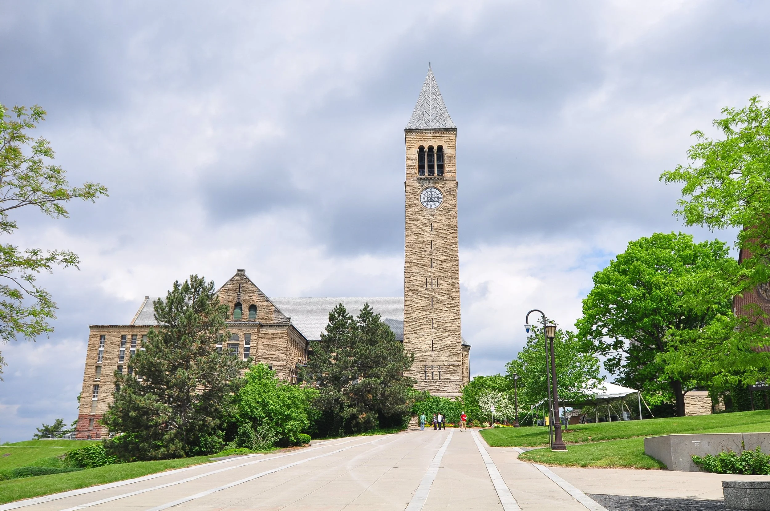 A historic stone campus church with a tall clock tower under a cloudy sky, surrounded by green trees and a paved walkway with a few people walking.