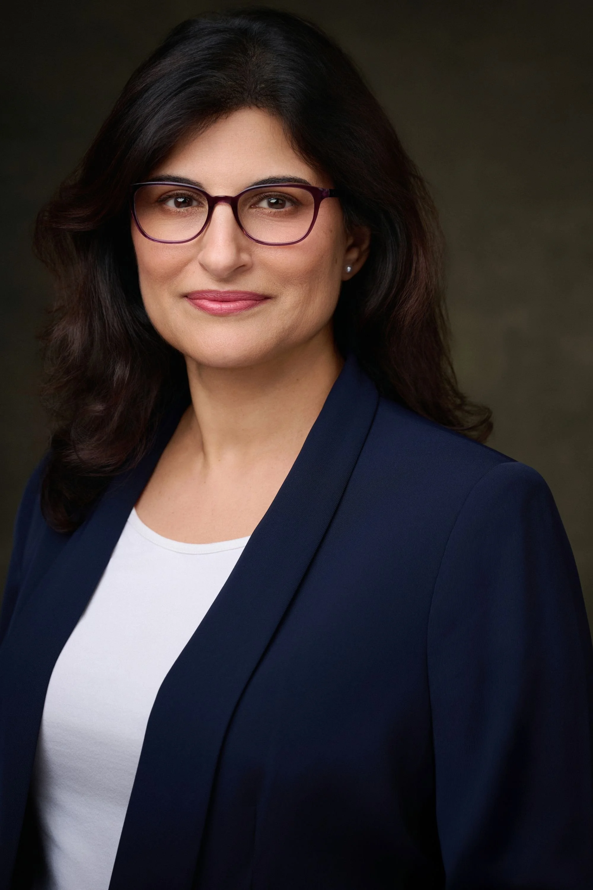 A professional woman with dark hair, wearing glasses, a navy blazer, and a white top, smiling with a dark, blurred background.