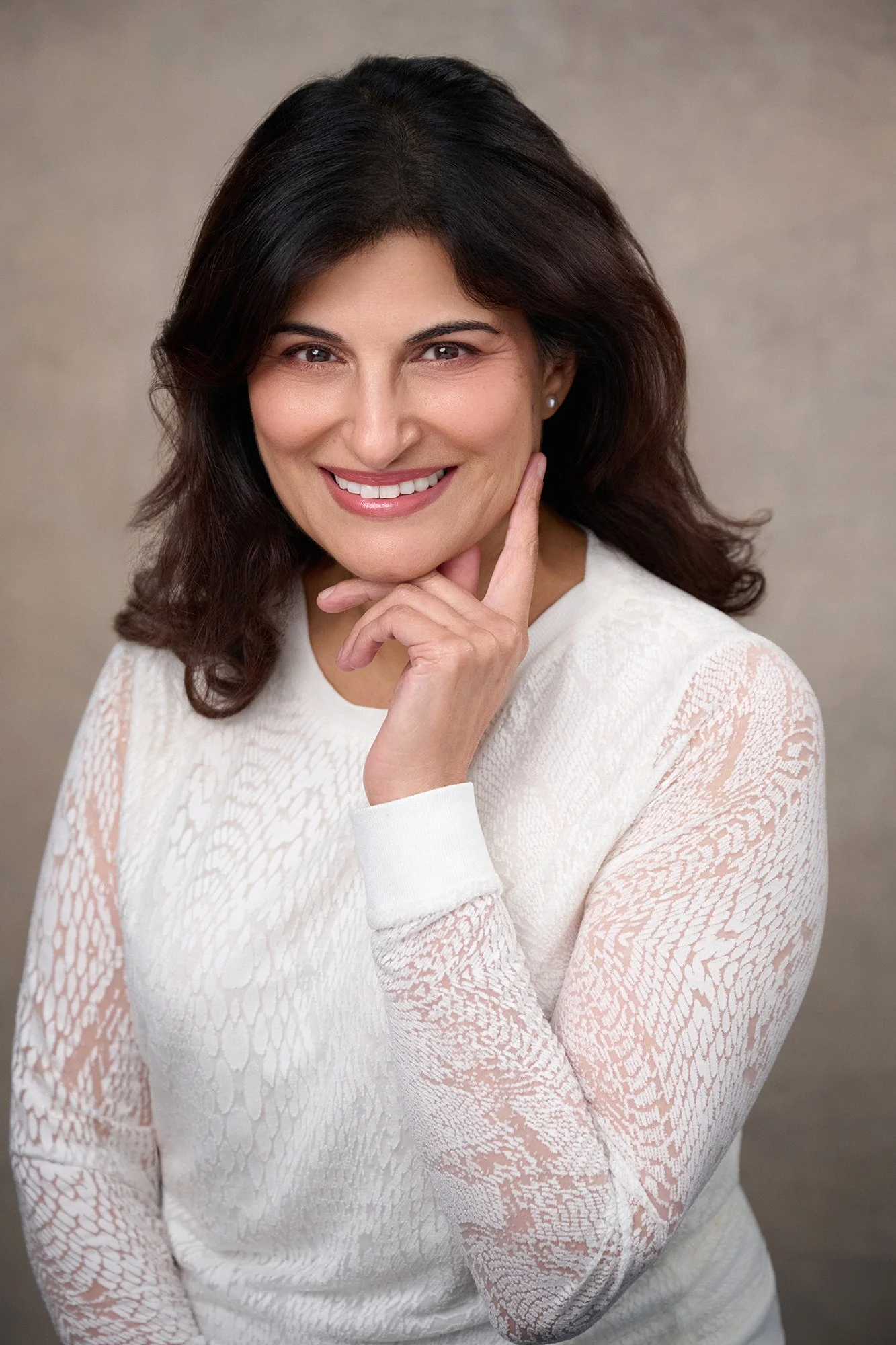 A woman with dark brown hair, wearing a white long-sleeve top with lace-like pattern, smiling and touch her chin with her left hand.