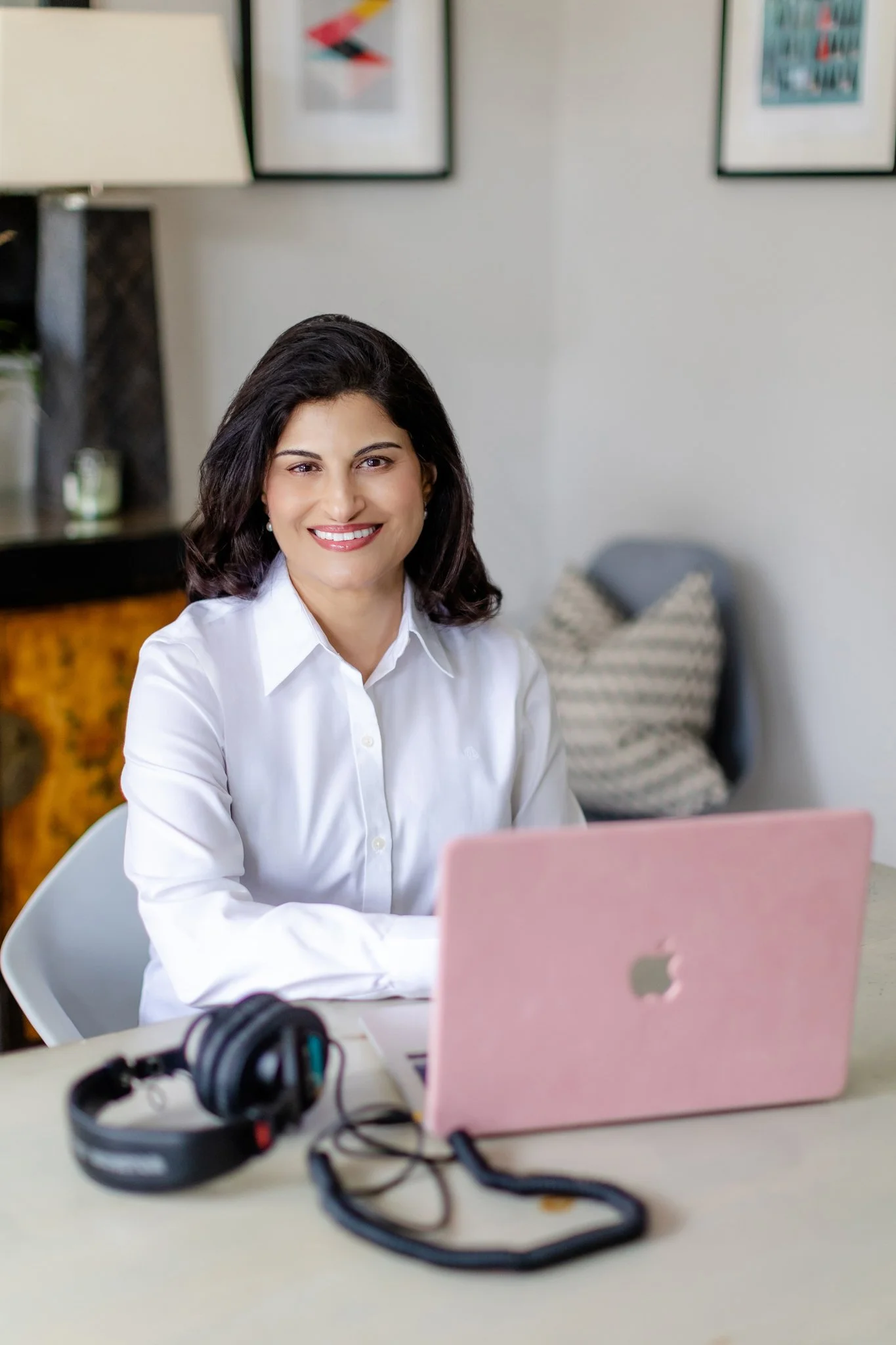 A woman smiling at her pink laptop during a video call or working from home, headphones on a beige table in front of her.