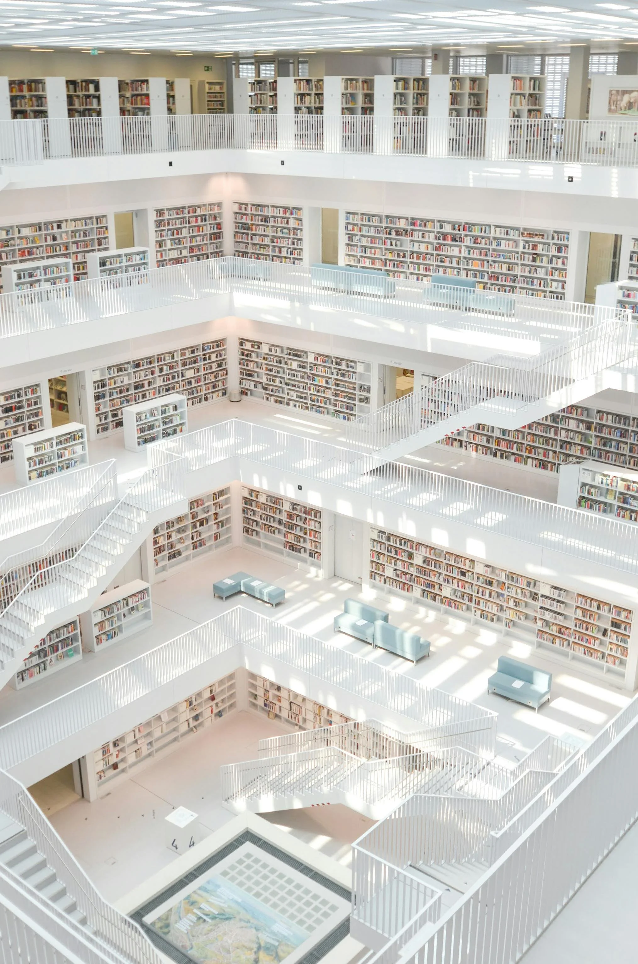 Interior of a bright, multi-story modern library with white shelves filled with books, white railings, and seating areas, illuminated by abundant natural light.