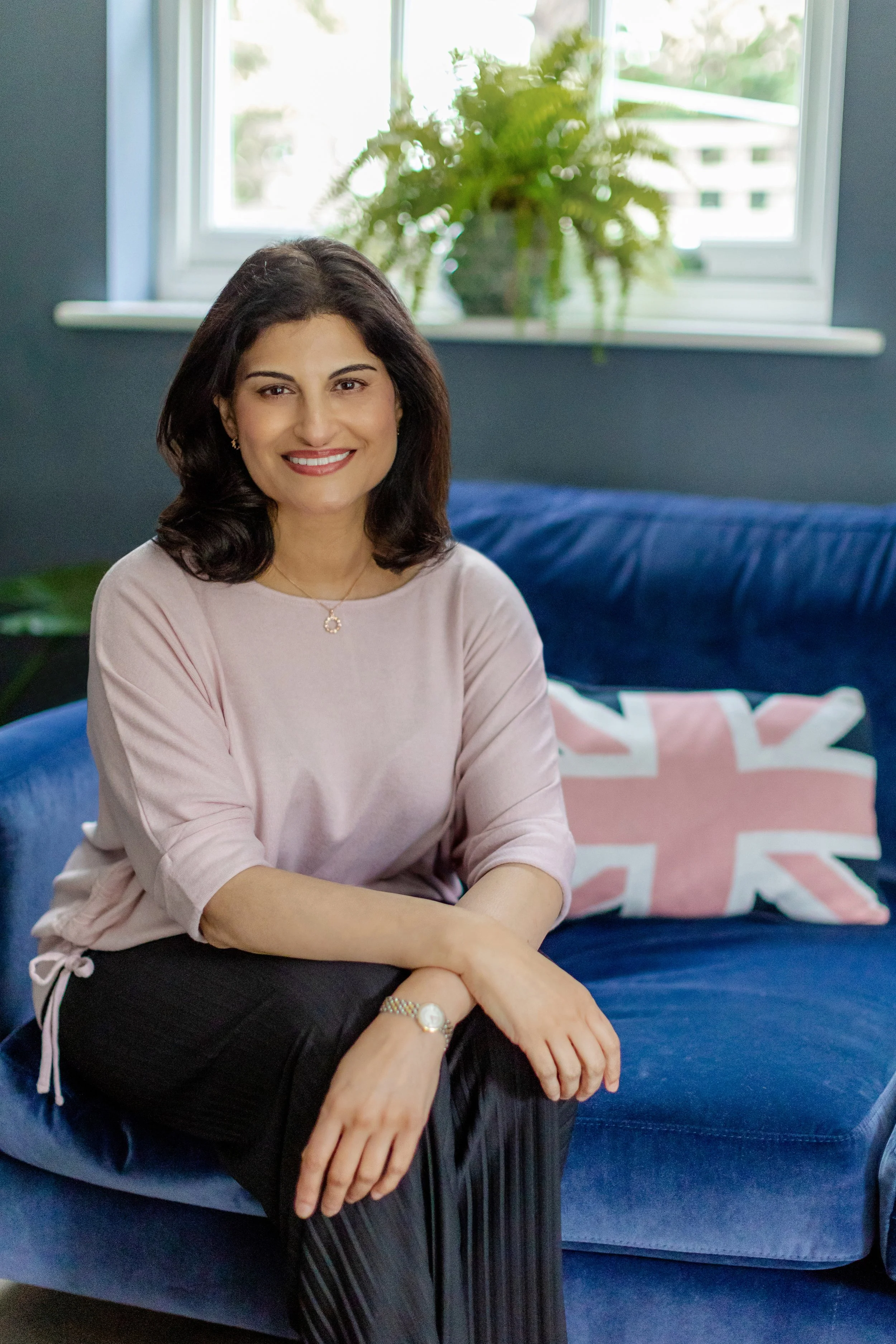 A woman with shoulder-length dark hair, wearing a light pink blouse and dark striped pants, sitting on a blue sofa with a Union Jack pillow, in a room with dark blue walls and a window with a potted plant in the background.